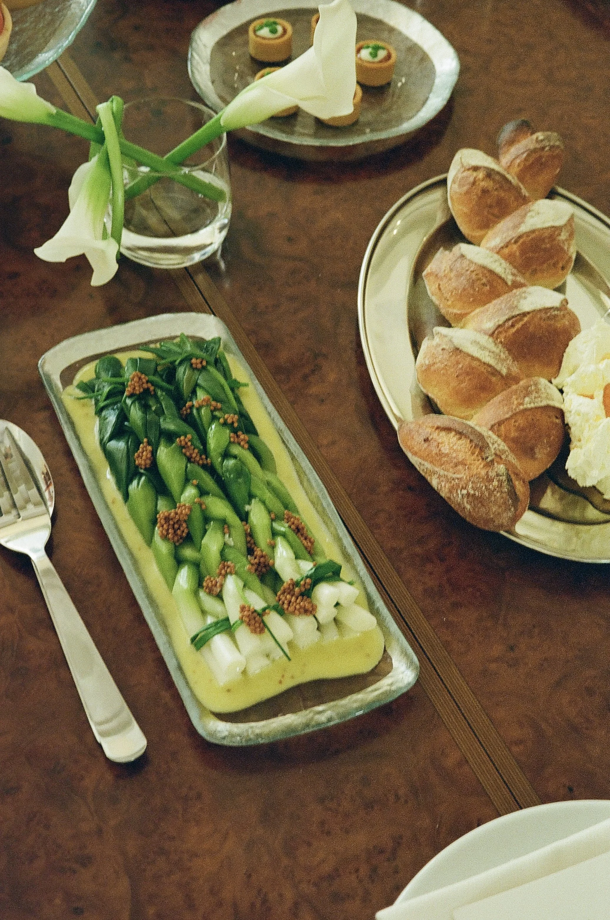 Een tafel met diverse gerechten, waaronder een schaal met groene groenten en witte knoflook, een bord met zachte broodjes en gerookte zalm, een glas met witte bloemen, en een bord met kleine sushirolletjes met groene toppings.