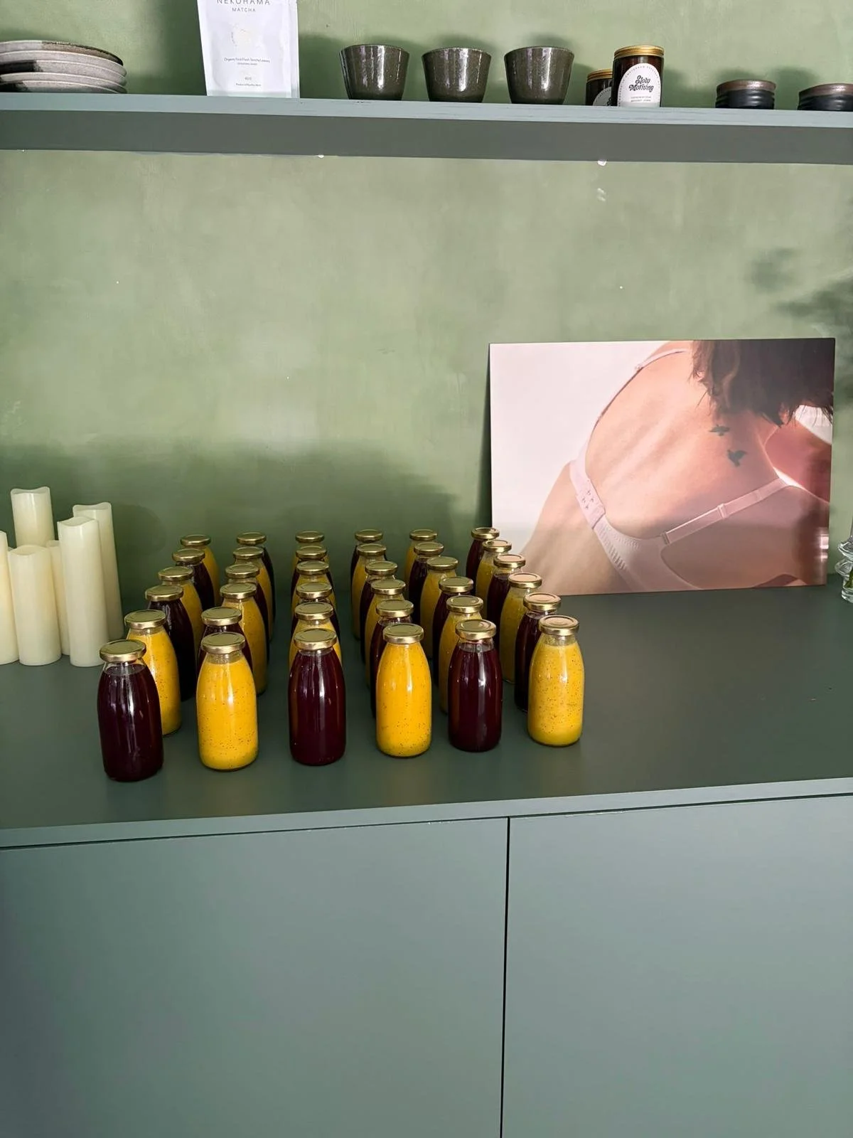 Een tafel met geprofileerde flessen, waarschijnlijk vruchtensap, in drie rijen. Op de achtergrond een foto van een vrouw met een tattoo in een BH of sporttop en witte straps, en een groene muur met opslagschalen en potjes.