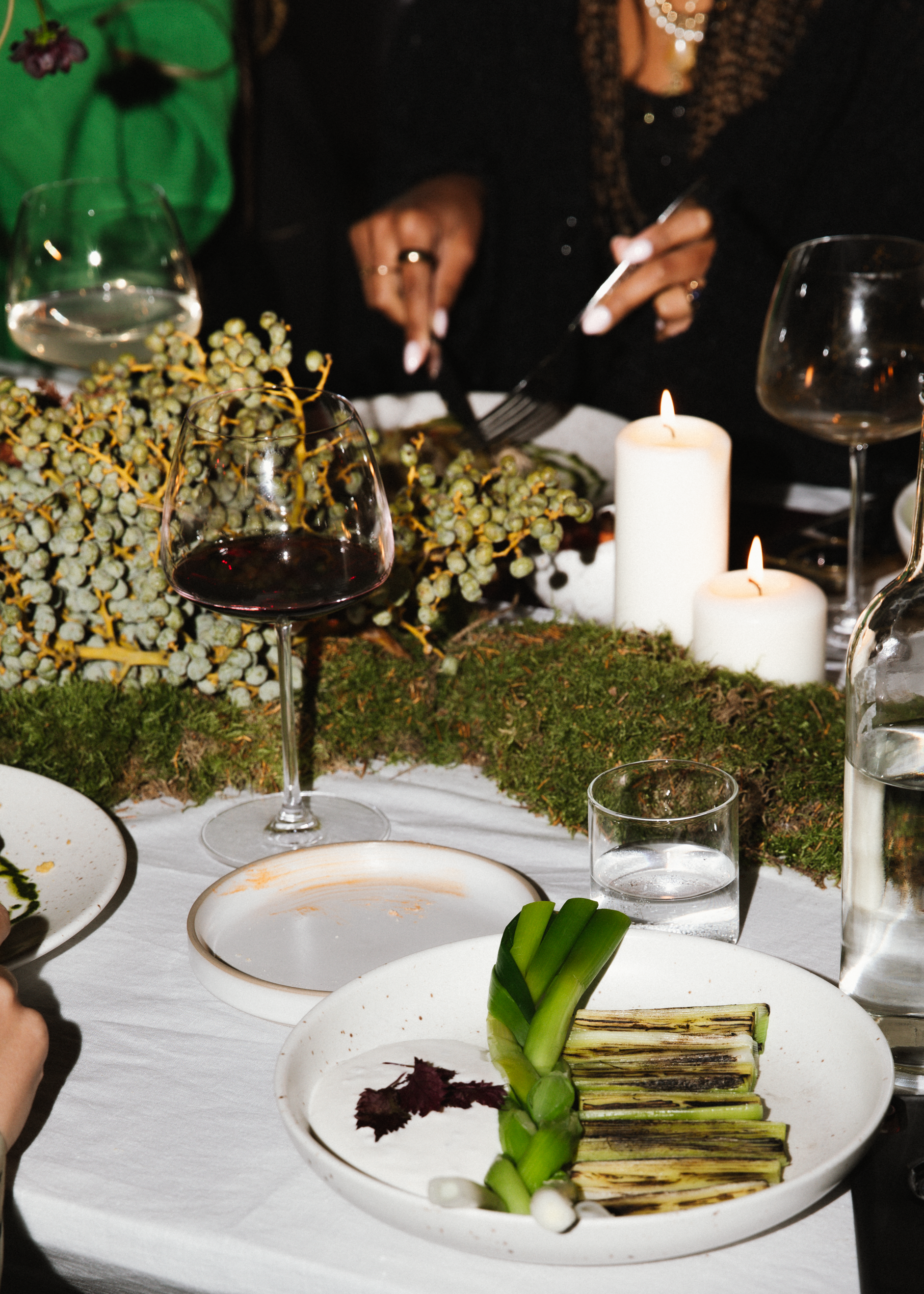 Een feesttafel met verschillende glazen, kaarsen, een bord met gegrilde asperges, en een bloemstuk met groene bladeren en witte bloemen.