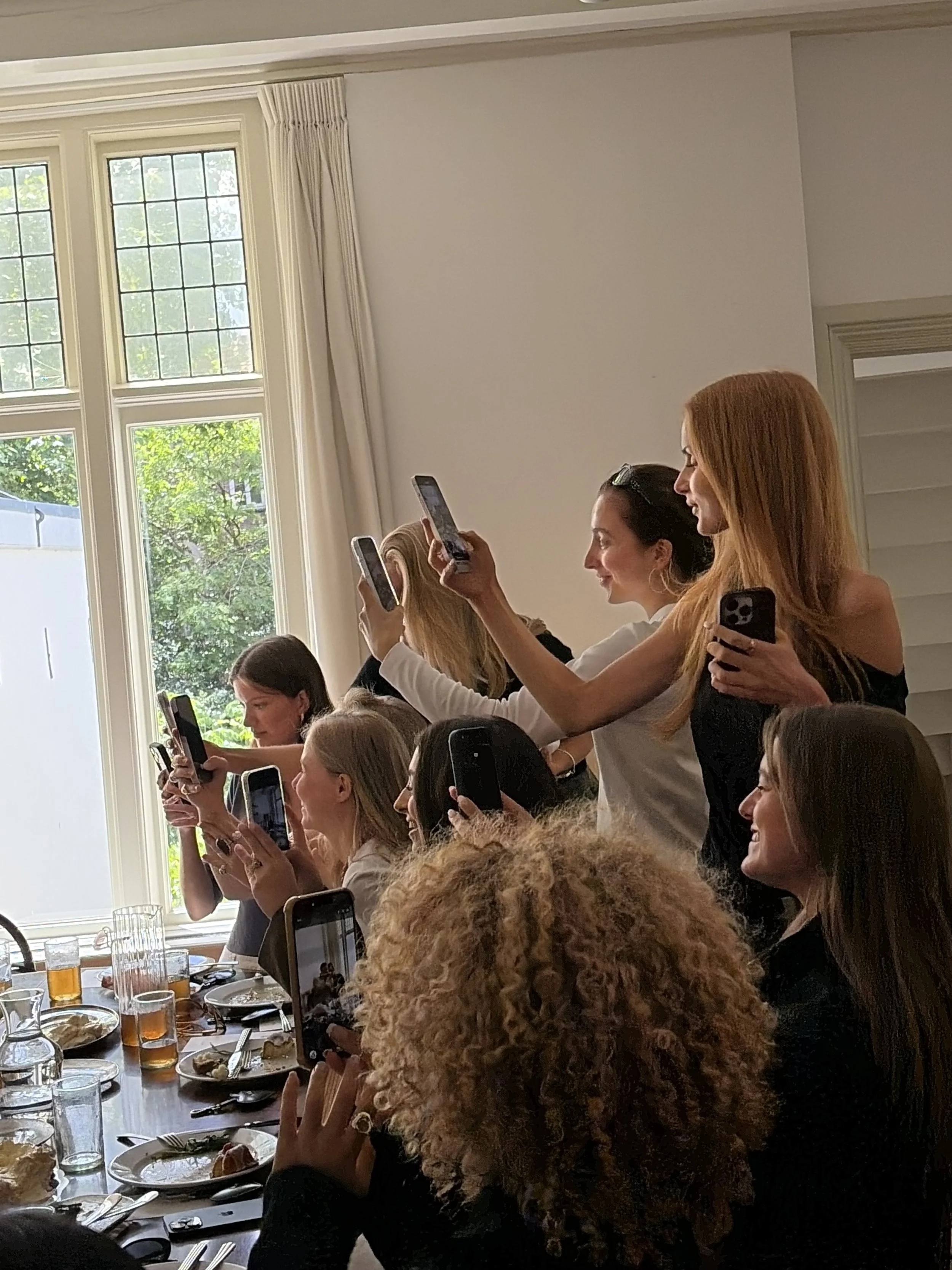 Vrouw met rood haar en een bril, die op haar telefoon foto's maakt tijdens een bijeenkomst met meerdere mensen, zittend aan een lange eettafel met drinken en eten.