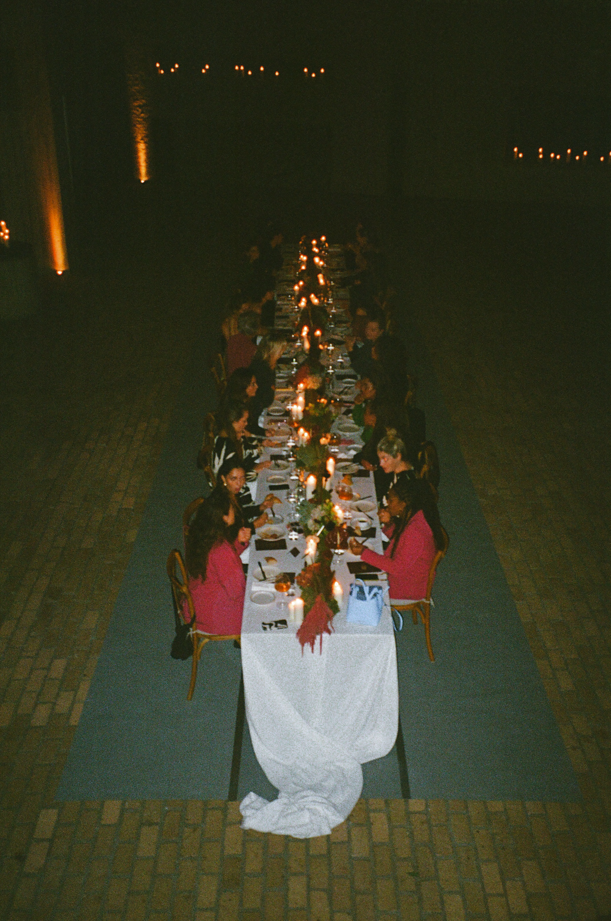Lange tafel met gasten tijdens diner, sommige gasten in roze outfits, kaarsen als verlichting, donker buiten.