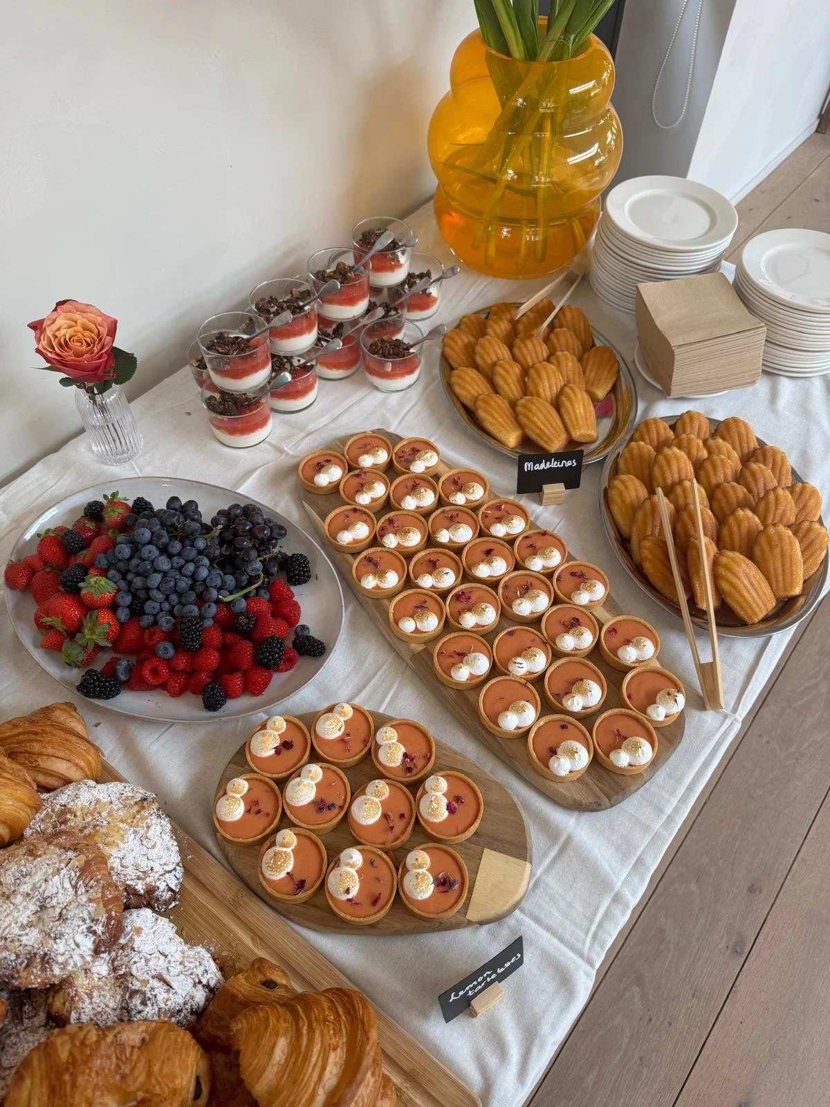 Een buffet met diverse desserts, vers fruit, smallereaying bakkerijtjes, een vaas met een bloem, en borden en papieren servetten.
