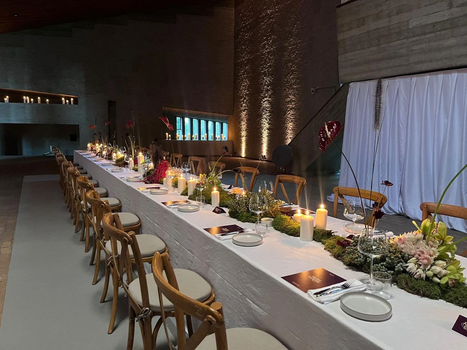 Een lang gedekte tafel met kaarsen, glazen en bloemen in een sfeervolle, moderne omgeving met houten muren.