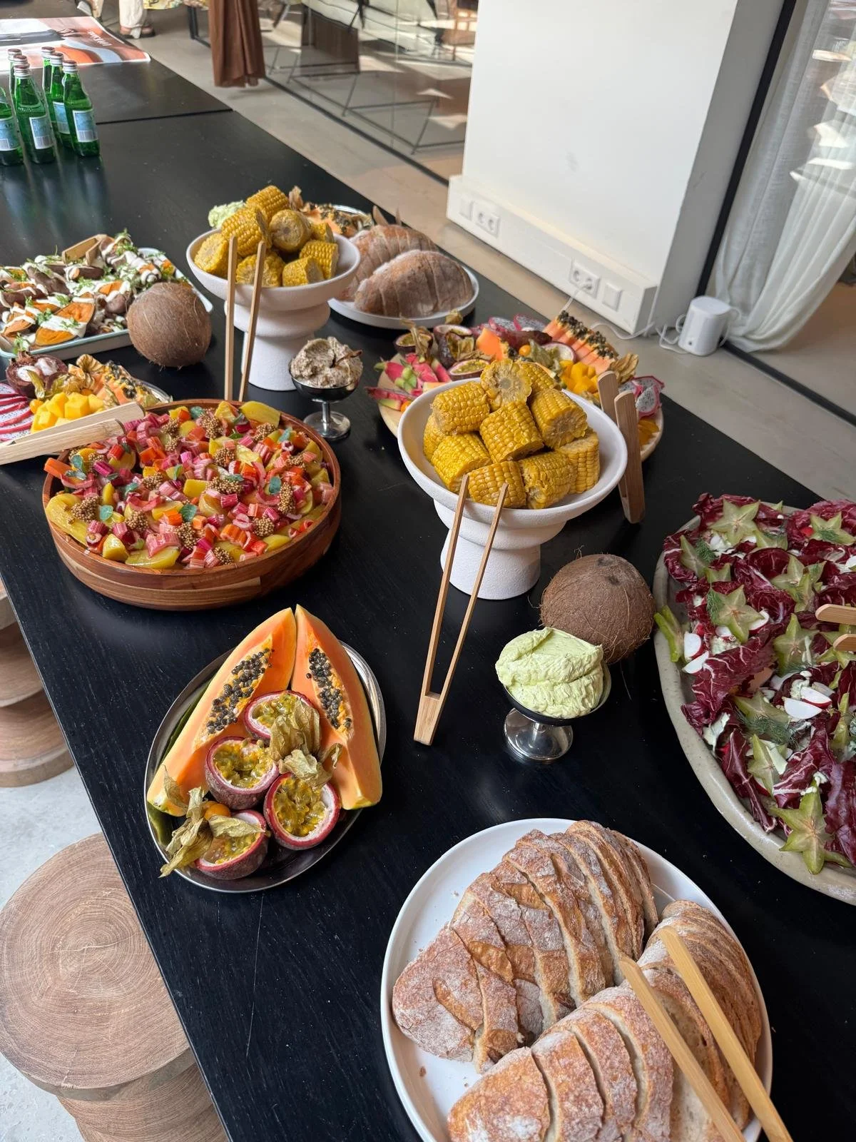 Een tafel met diverse gerechten, waaronder maïskolven, fruit, salade, brood en andere hapjes, klaar voor een feestbuffet.