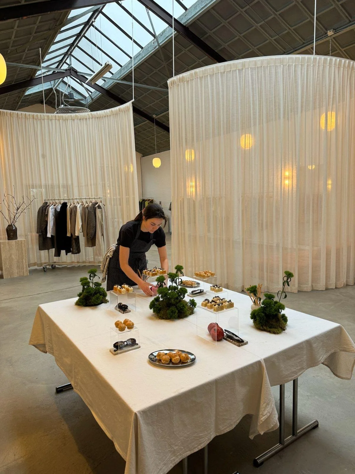 Vrouw serveert kleine desserts op een wit tafelkleed, omgeven door groene decoratie, binnen in een moderne winkel of tentoonstelling met gordijnen en kleding in de achtergrond.