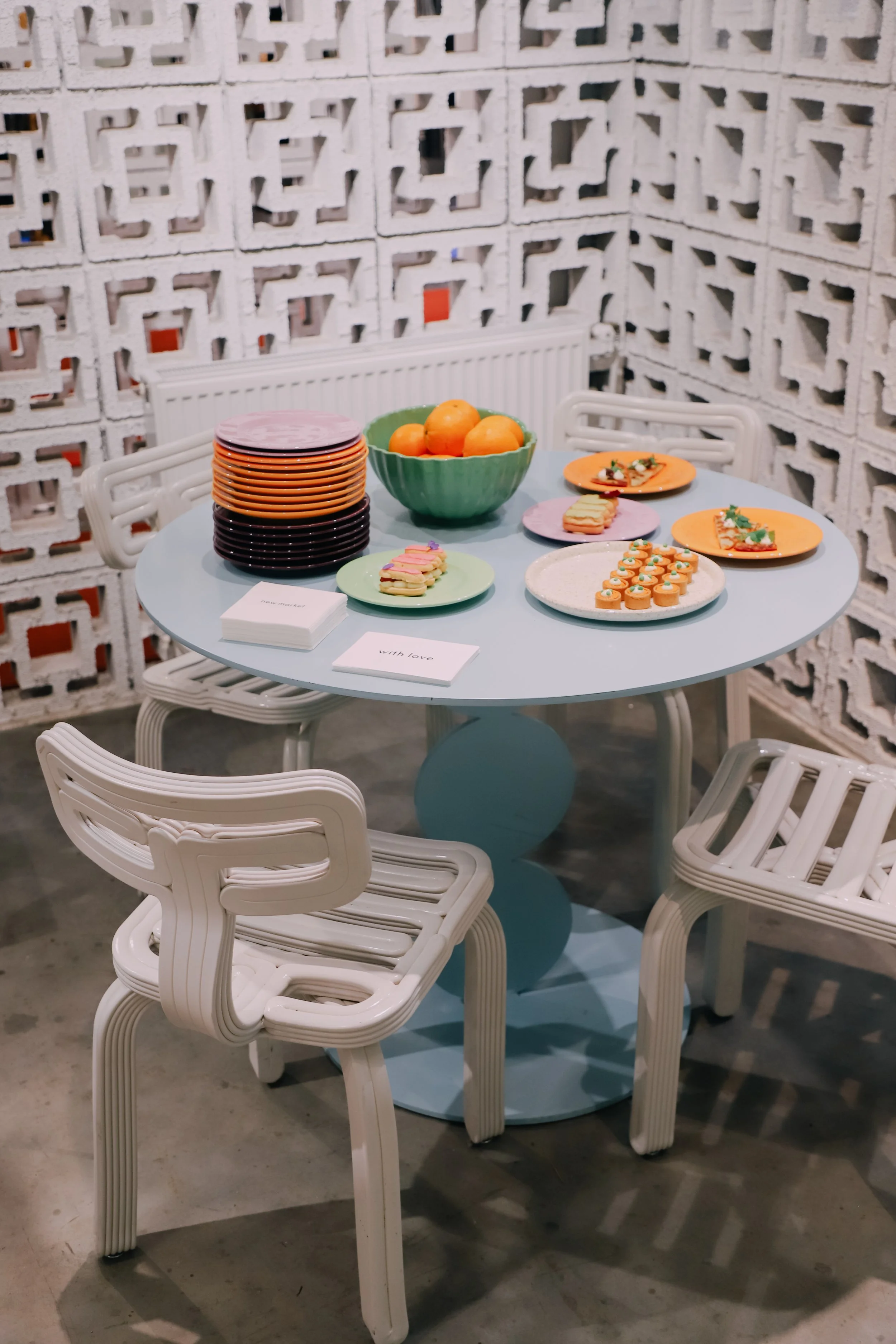 Een ronde tafel met witte plastieken stoelen eromheen. Op de tafel liggen verschillende kleurrijke borden met hapjes, een kom met oranje mandarijnen, en een stapel kleurrijke borden. Op de tafel liggen ook servetten met de tekst 'with love'. De achte