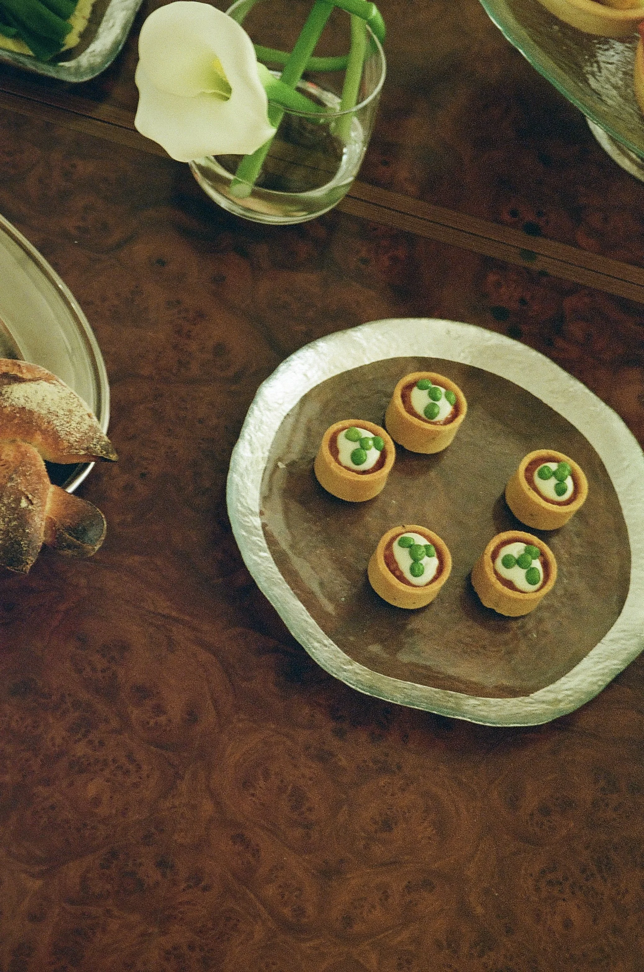 Mini taartjes met groene decoratie op een zilveren schaal, een glas met een witte bloem, een stuk brood en een bord met een artikel met groene decoraties op een houten tafel.