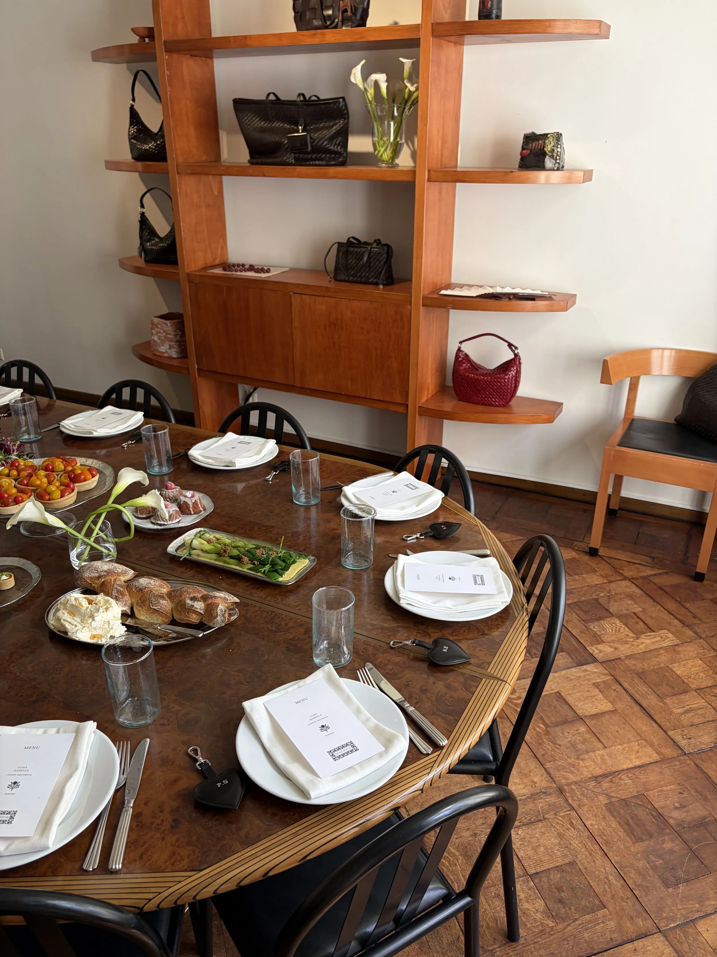 Een eetkamer met een grote houten tafel, klaargemaakt voor een maaltijd met servetten, borden, bestek, glazen en menu's. Op de tafel liggen ook verschillende gerechten en desserts. Achter de tafel staat een houten boekenkast met handtassen en decorat