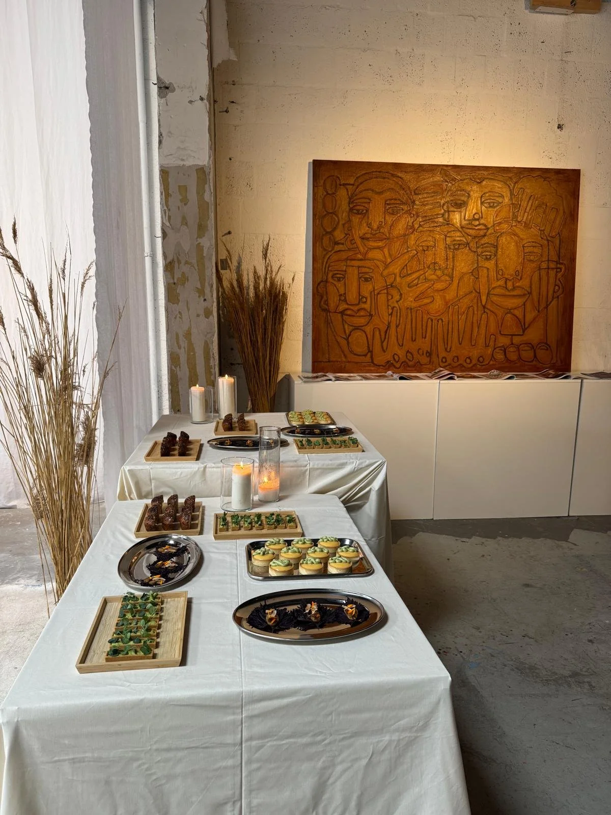Een tafel met diverse hapjes en desserts, versierd met kaarsen, in een interieur met een abstract kunstwerk aan de muur en decoratieve droge planten.