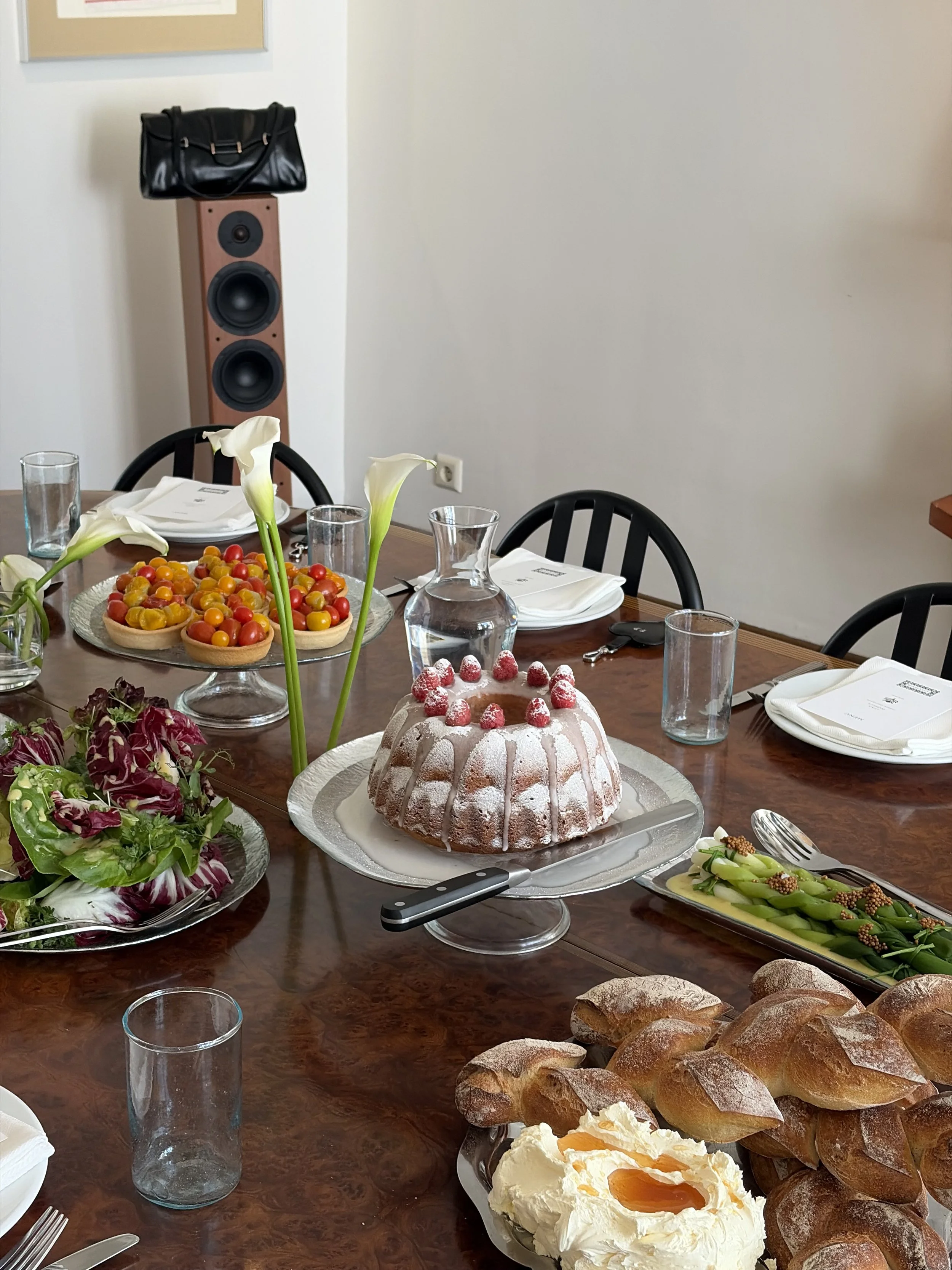 Een gedekte eettafel met brood, een gebak met aardbeien, een fruitsalade, bloemen, glazen water, borden en bestek, op de achtergrond een luidspreker en een zwart handtas.