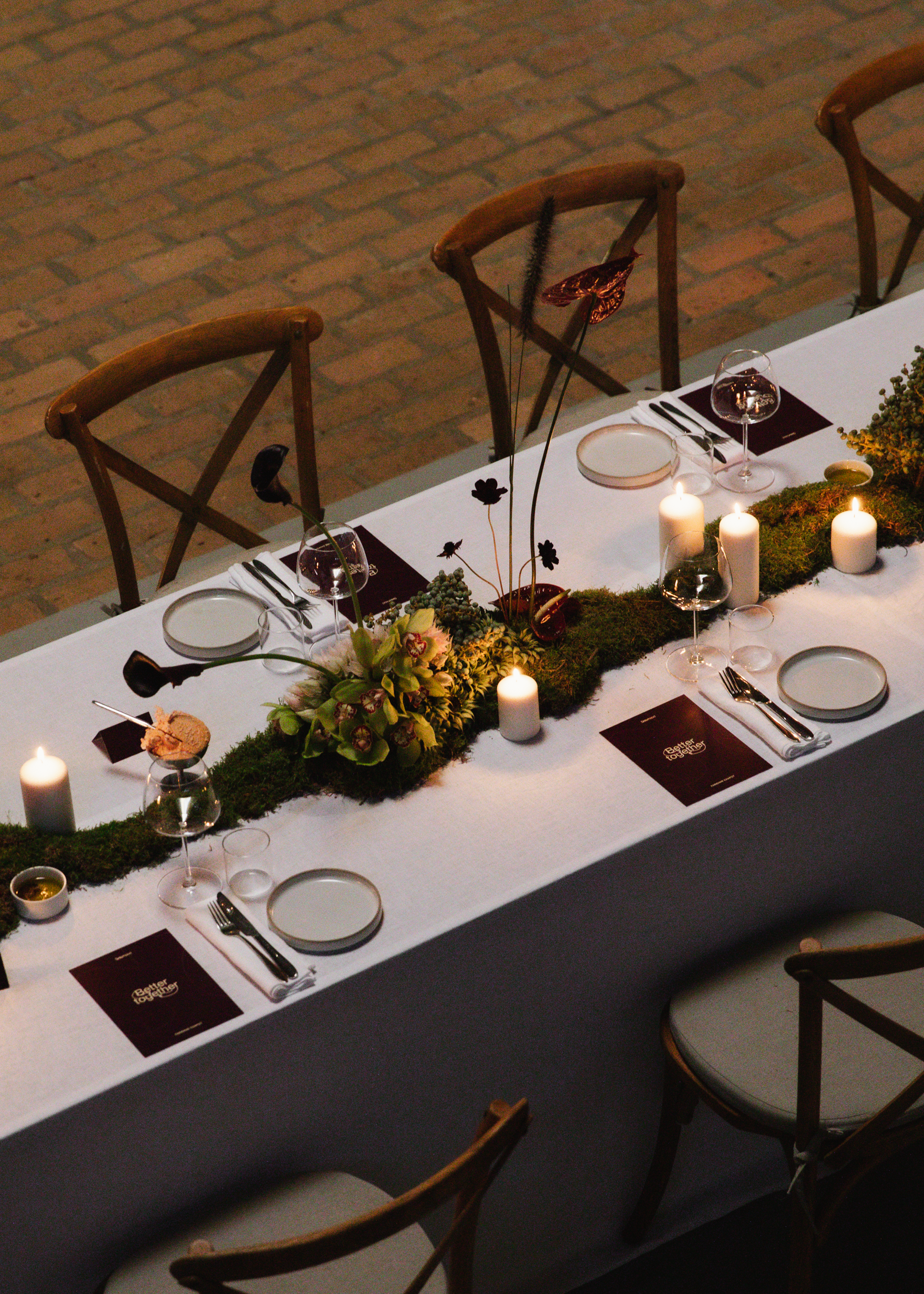 Een lange feesttafel met witte tafelkleed, kerstmutsen, kaarsen, glazen wijn, borden, bestek, en bloemen in het midden; houten stoelen rondom. Smalle strook bloementak versierd. Op de tafel liggen menu-kaarten met teksten 'Eetter Together'.
