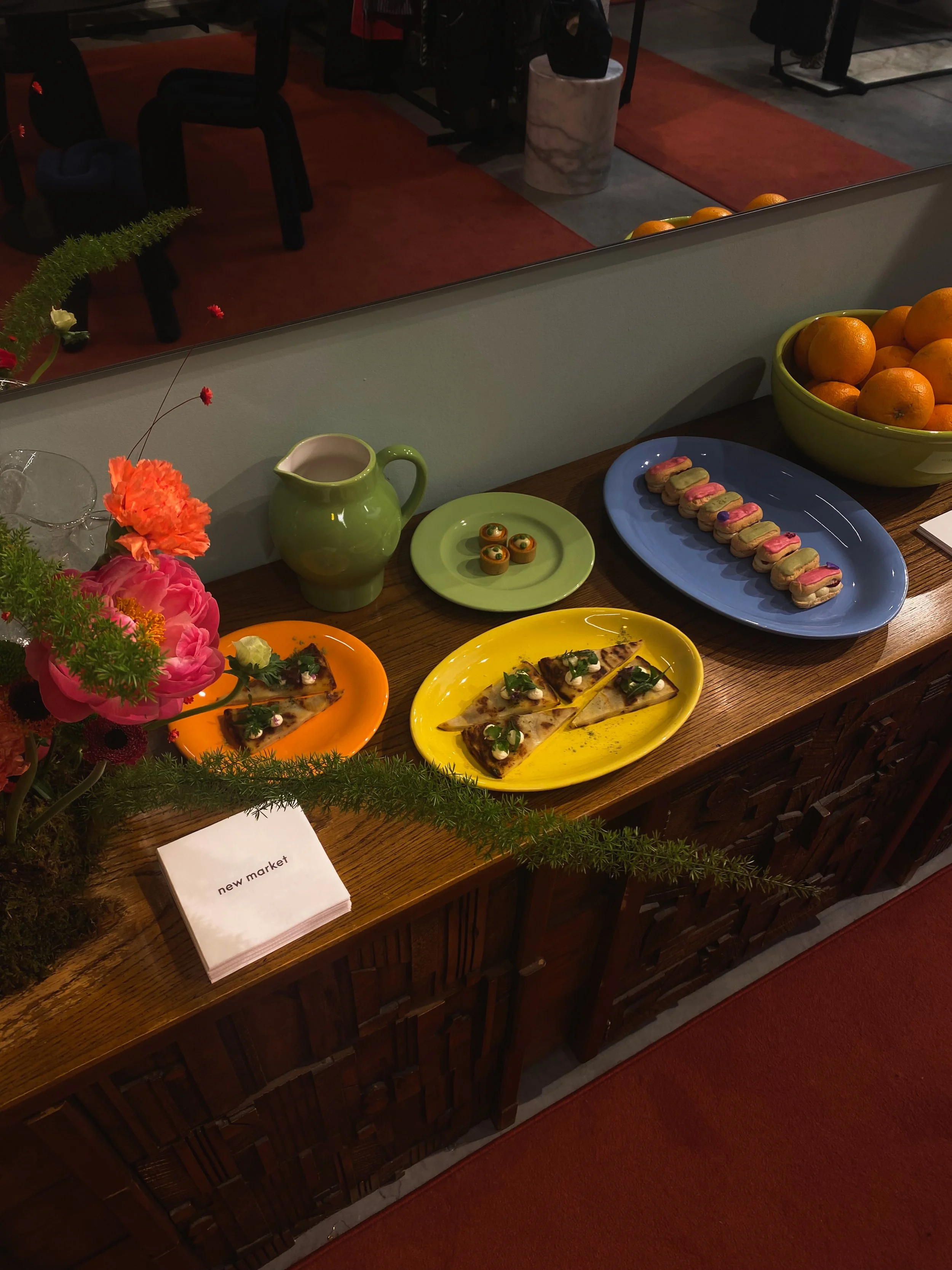Een tafel met verschillende kleurrijke gerechten, een vaas met bloemen, een komteneut met oranje's, en een bord met kleine taartjes, op de achtergrond een spiegel en stoelen te zien.