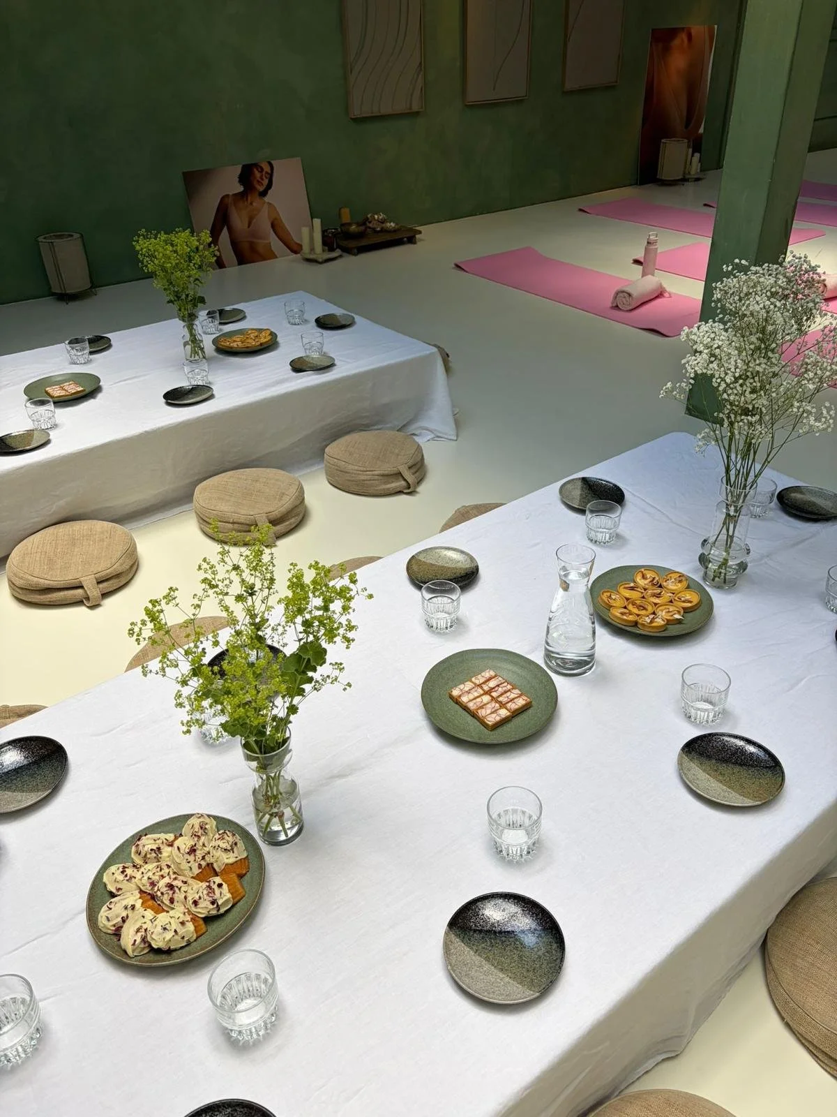 Gezellige eetruimte met langwerpige tafel, bedekt met een witte tafelkleed, met vazen met bloemen, serveerplaten met snacks, waterglazen, en kleine borden. In de achtergrond ligt een yogaruimte met matten, handdoeken, en een poster van een vrouw.