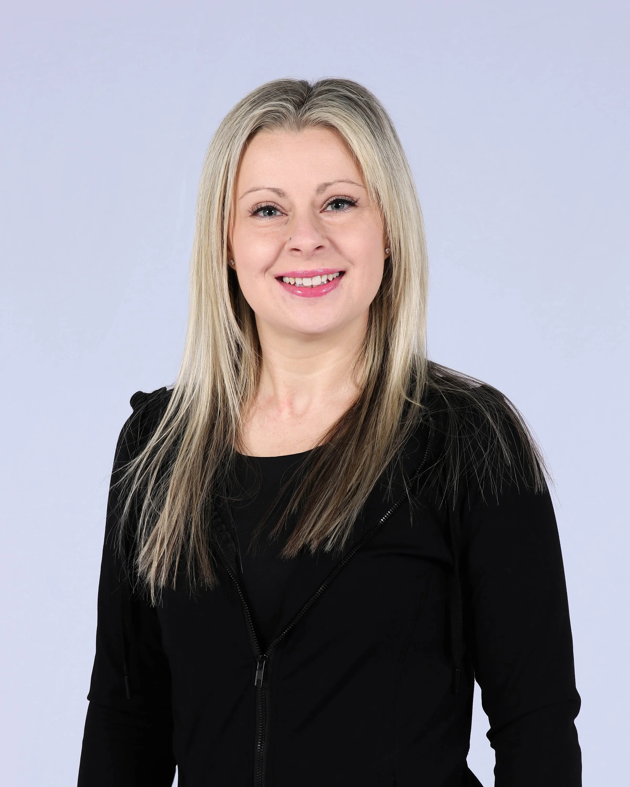 A woman with long blonde hair wearing a black top and jacket, smiling against a plain light background.