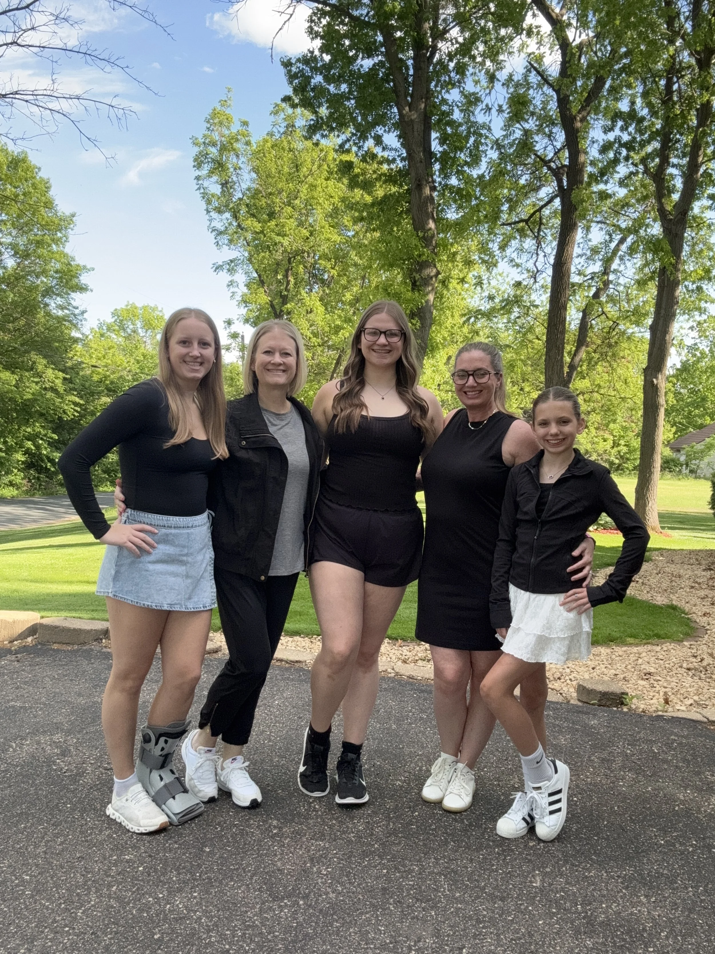 Five women standing outdoors on a paved area, smiling, with green trees and grass in the background.