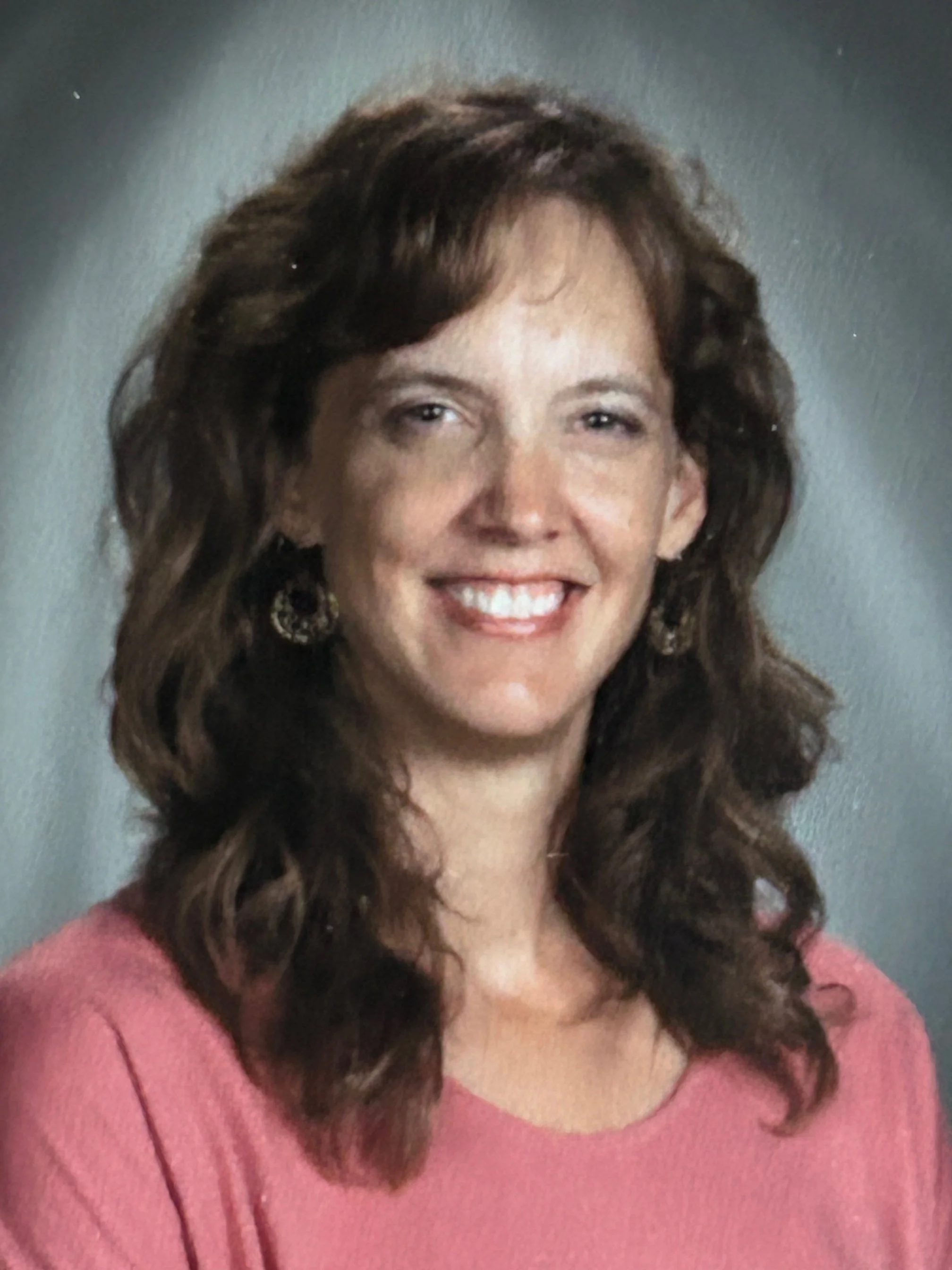 A woman with shoulder-length wavy brown hair, smiling, wearing a pink top and earrings, posed against a gray background.