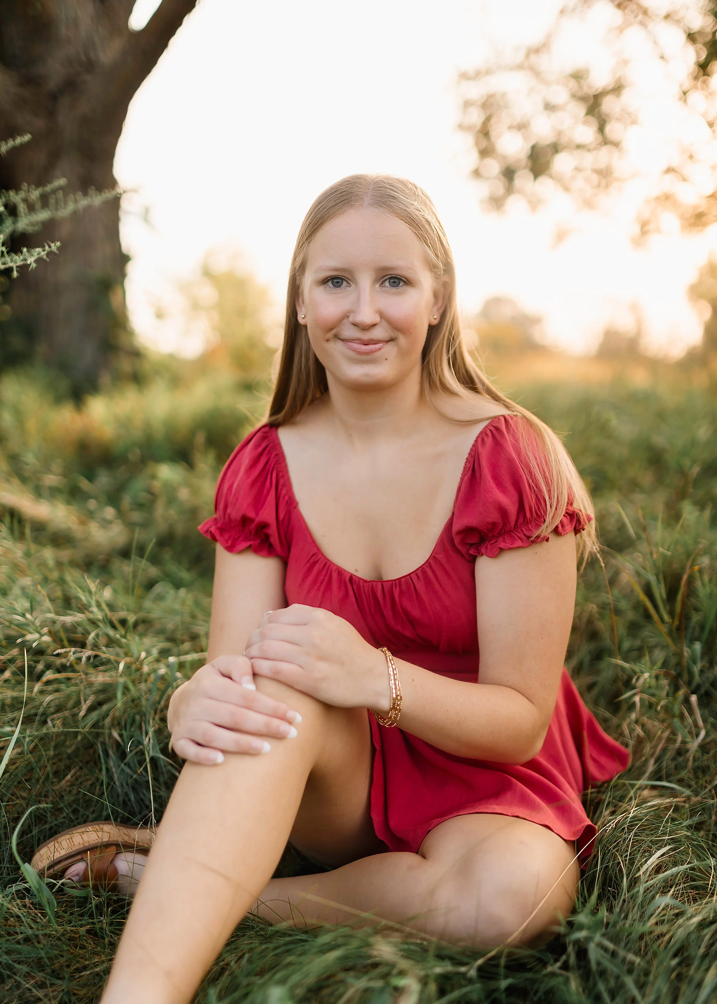 A young woman with long blonde hair in a red dress sitting on grass outdoors during sunset, smiling at the camera.