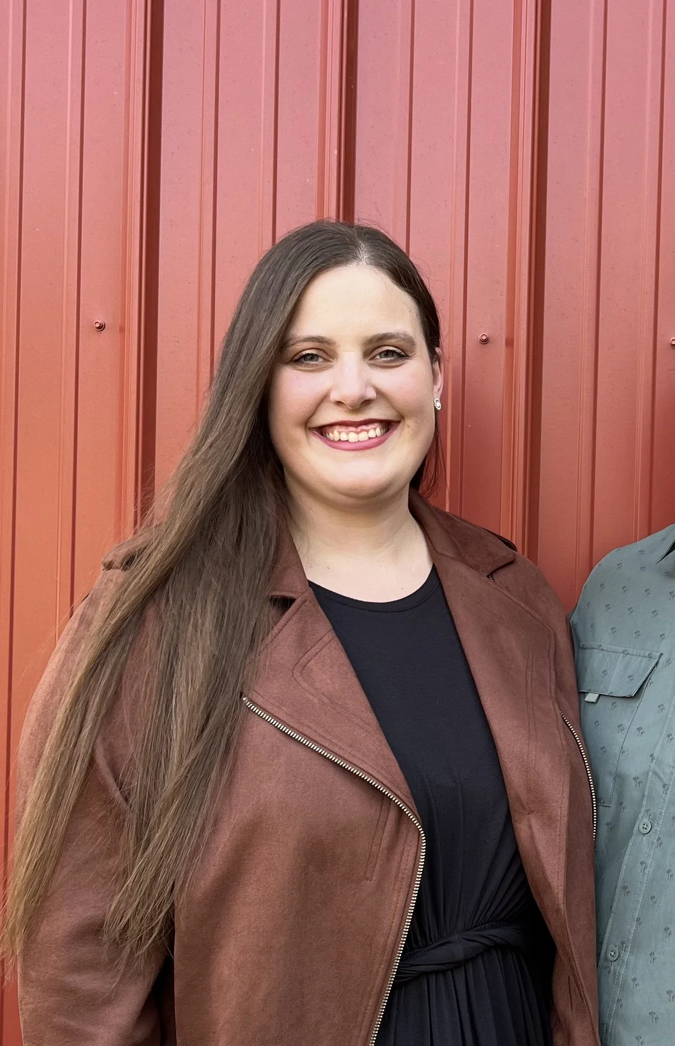 A woman with long brown hair and fair skin smiling in front of a red metal wall, wearing a brown jacket over a black top.