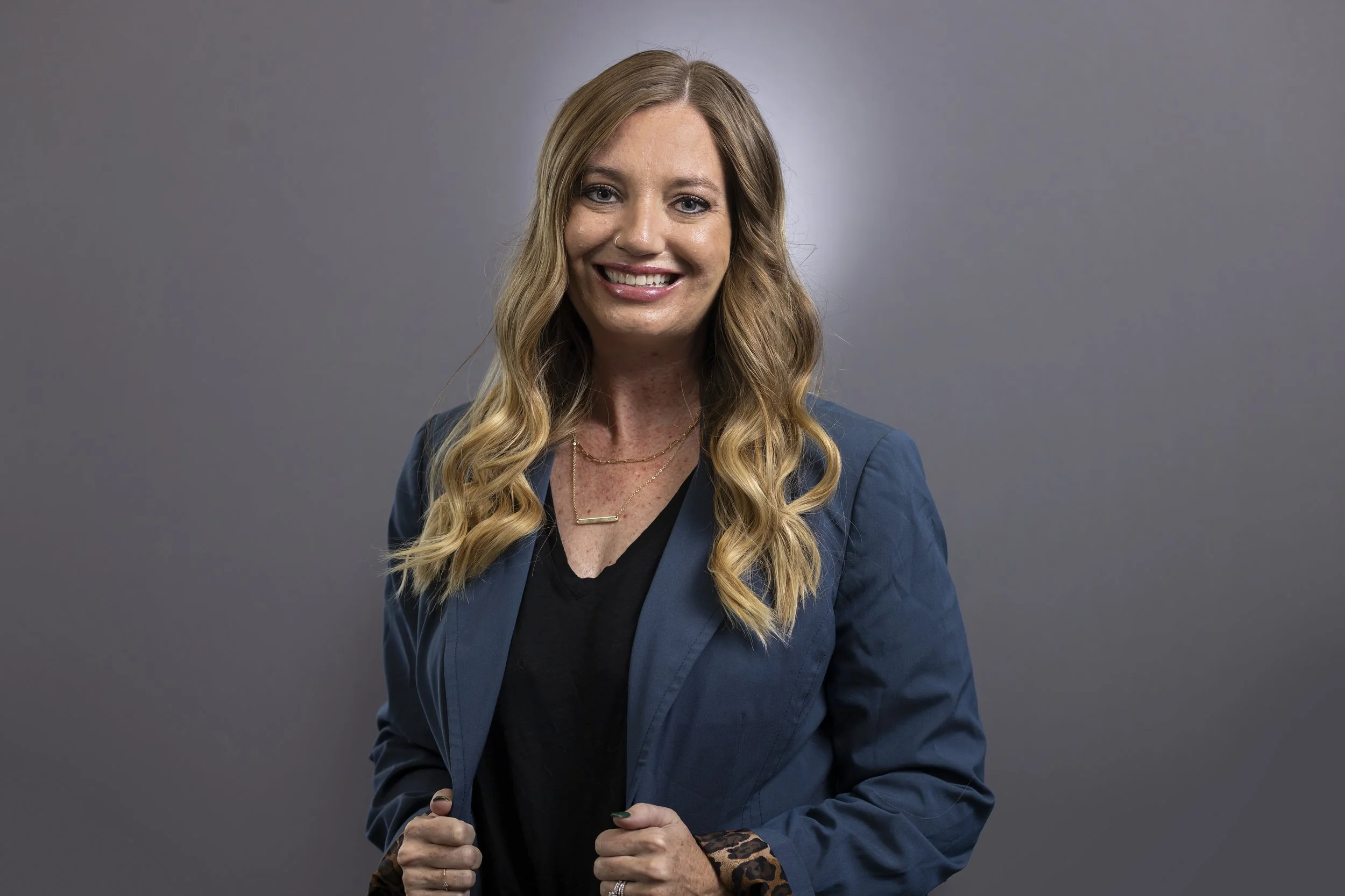 A woman with long wavy blonde hair in a navy blazer smiling at the camera, standing against a plain gray background.