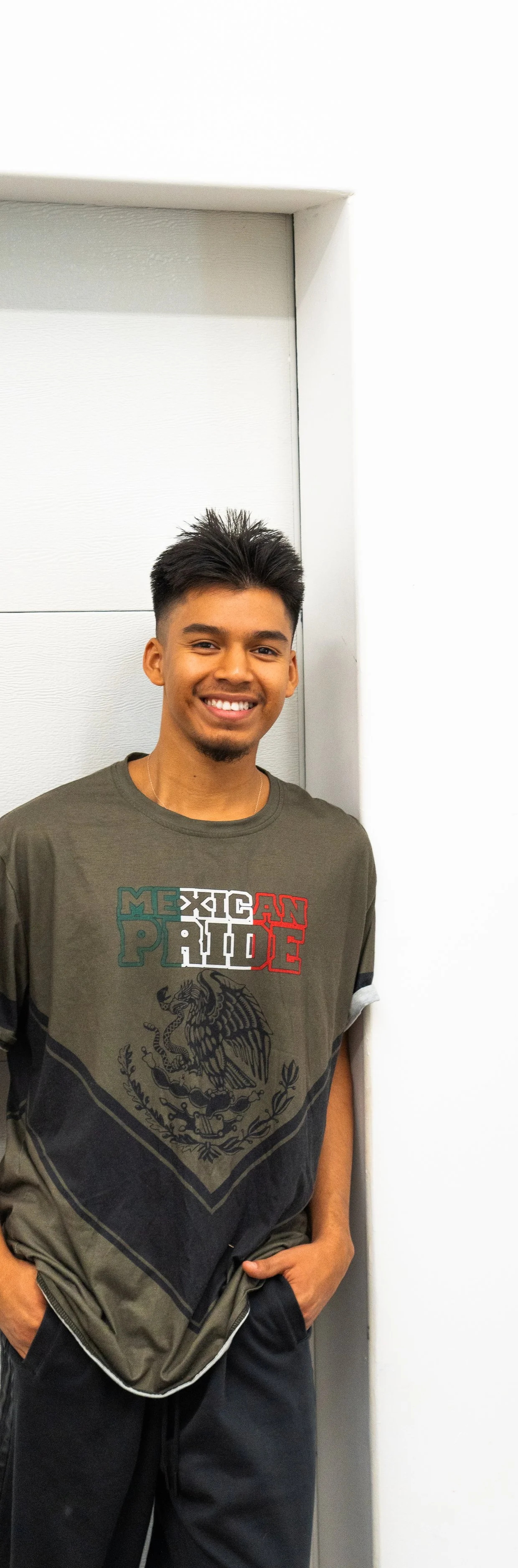 A young man with dark hair, smiling, standing against a white wall, wearing a gray T-shirt with Mexican pride text and a dragon emblem.