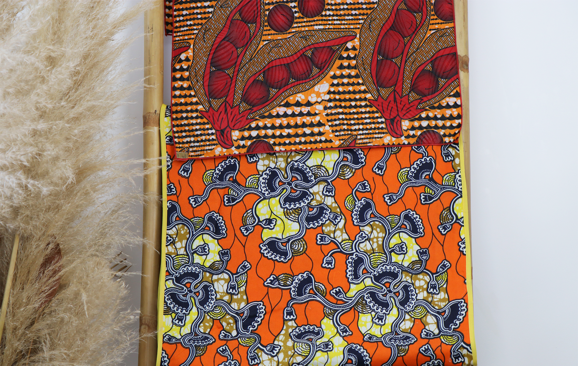 Deux morceaux de tissu aux motifs colorés, l'un avec des motifs rouges et oranges, l'autre avec des motifs floraux noirs sur fond orange, accrochés à une structure en bambou. Plantes séchées sur la gauche.