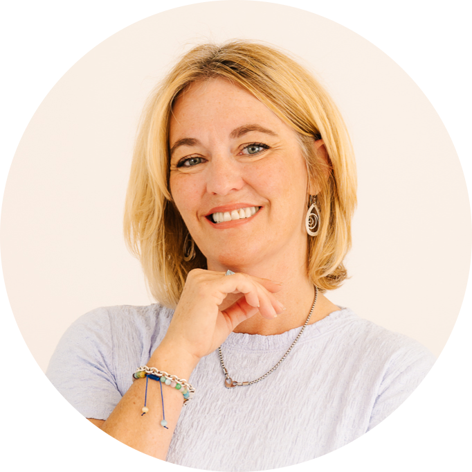 A smiling woman with blonde hair wearing a gray blazer and white shirt, sitting indoors.