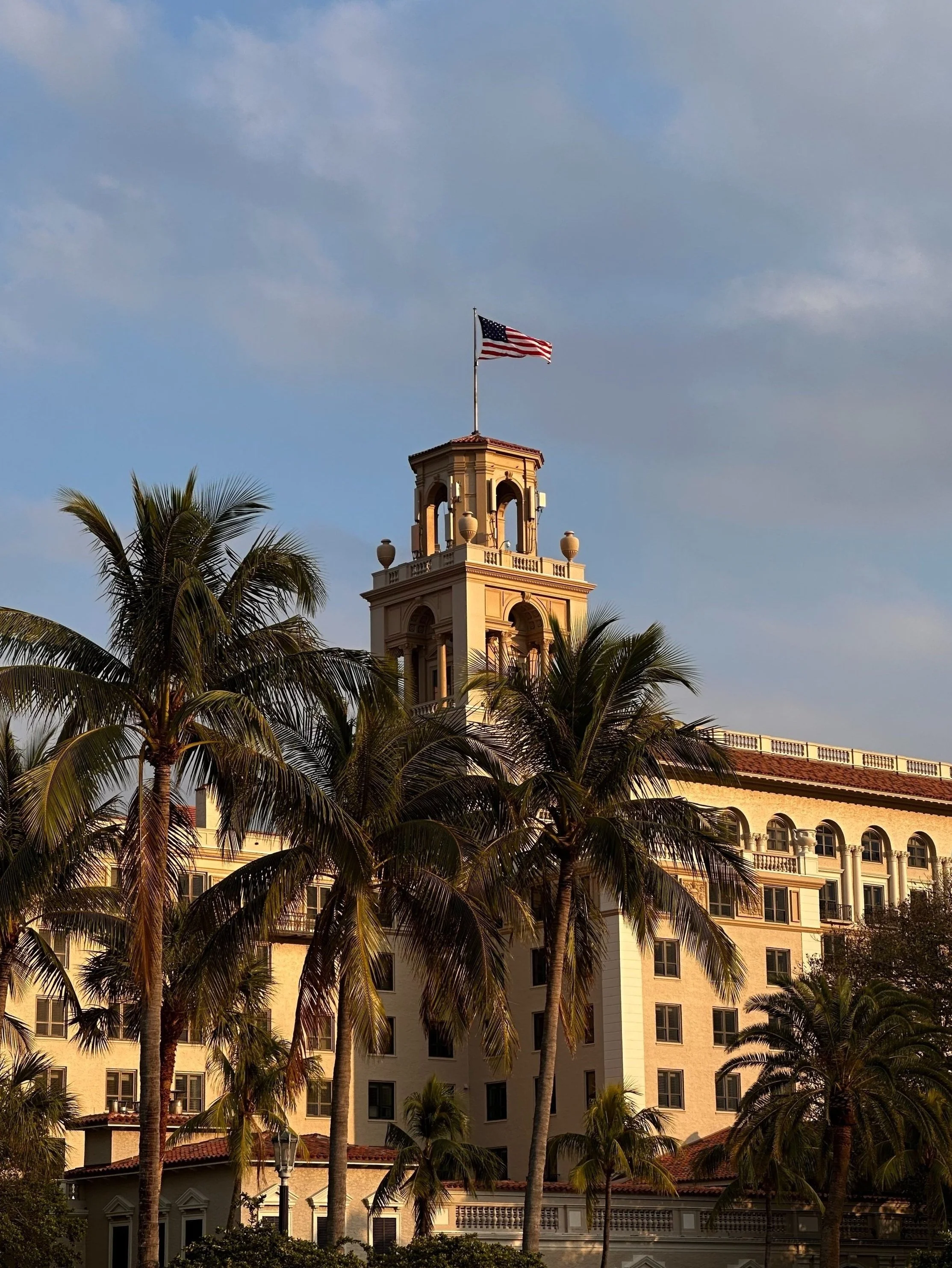 A historic building with a tower and an American flag, surrounded by palm trees, under a partly cloudy sky.