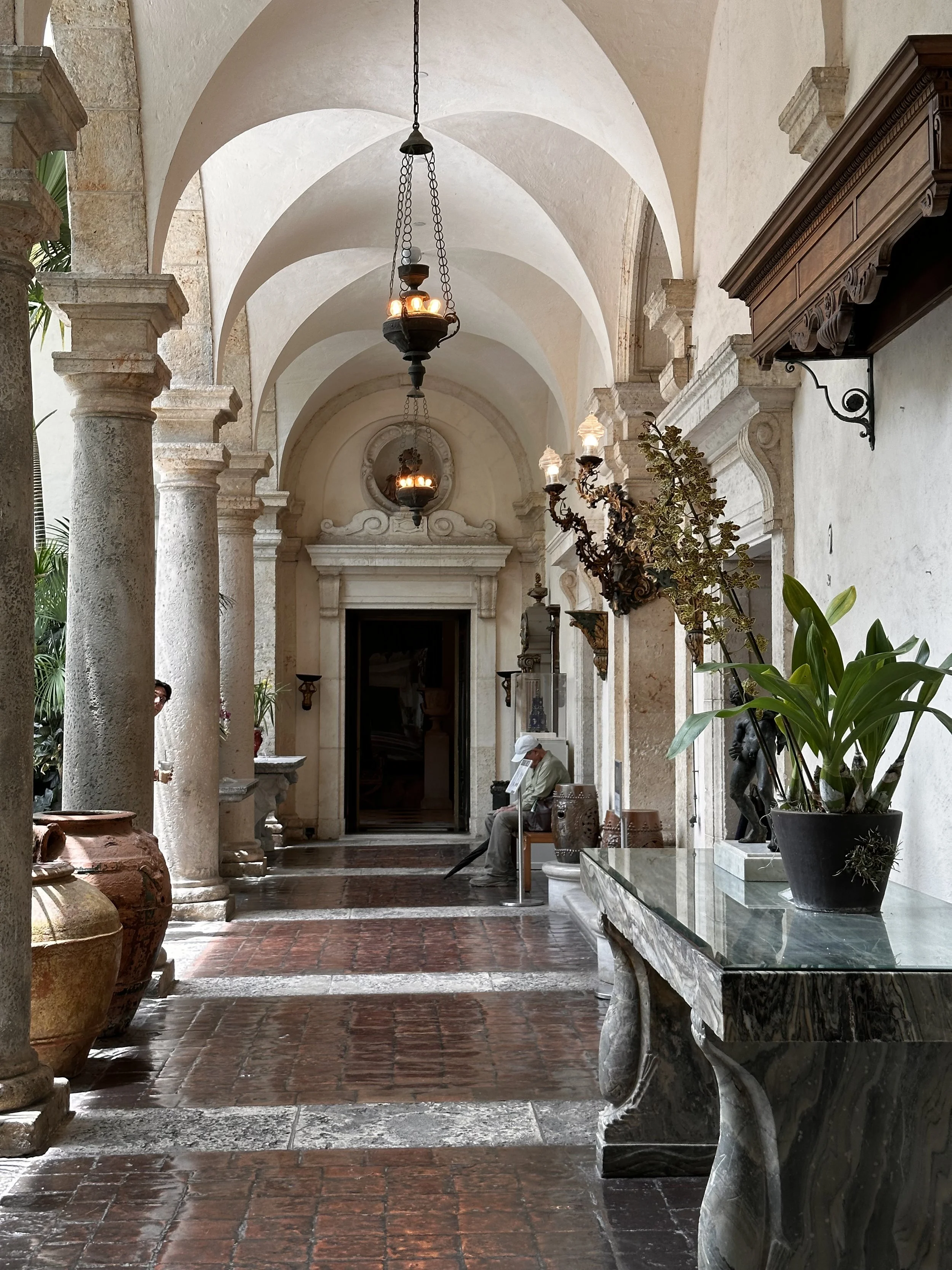 Indoor corridor with stone columns, hanging chandeliers, potted plants, and a person sitting on a bench.
