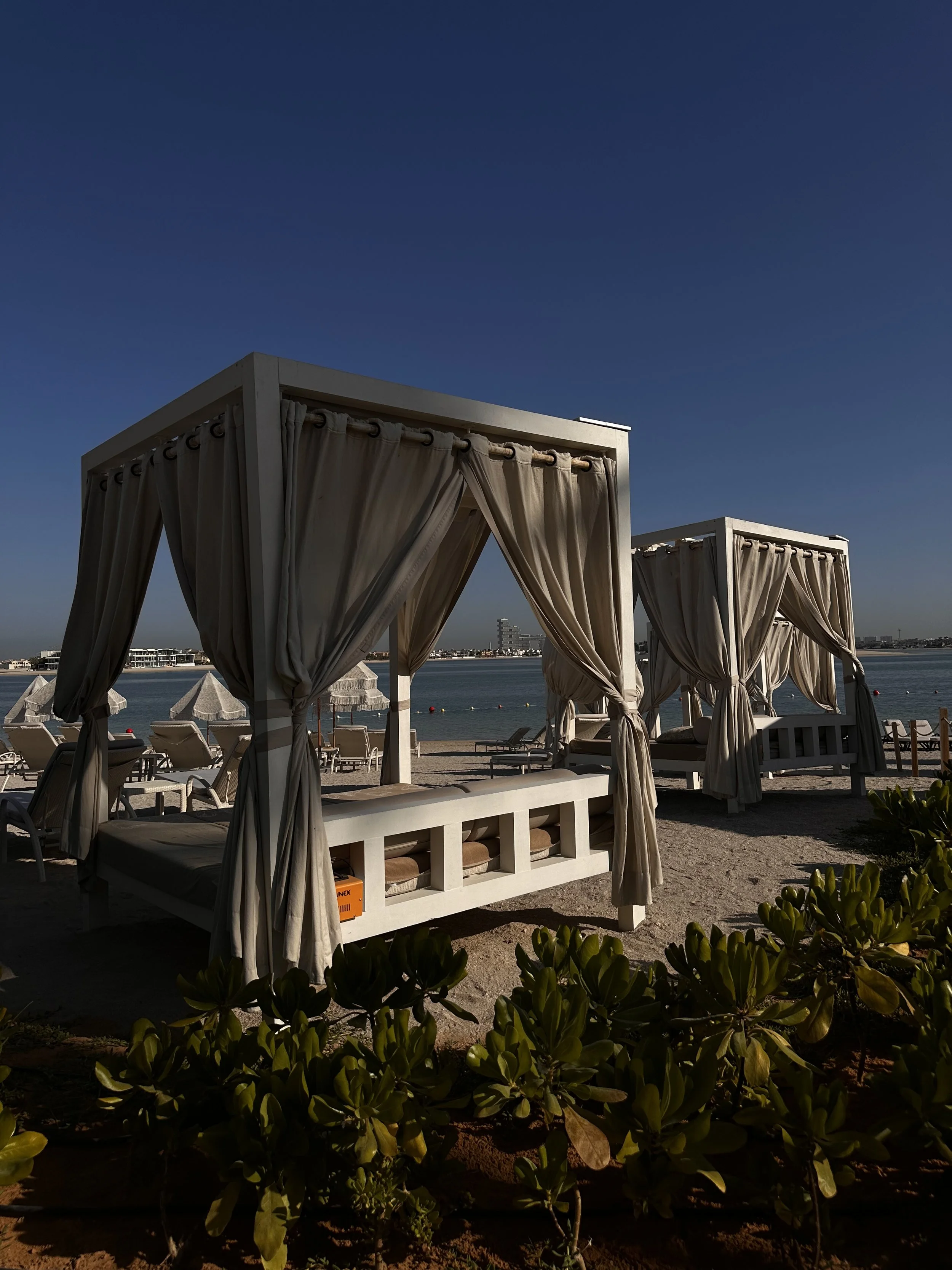 Beach with two white cabanas with curtains, lounge chairs, and umbrellas, overlooking calm water under a clear blue sky.
