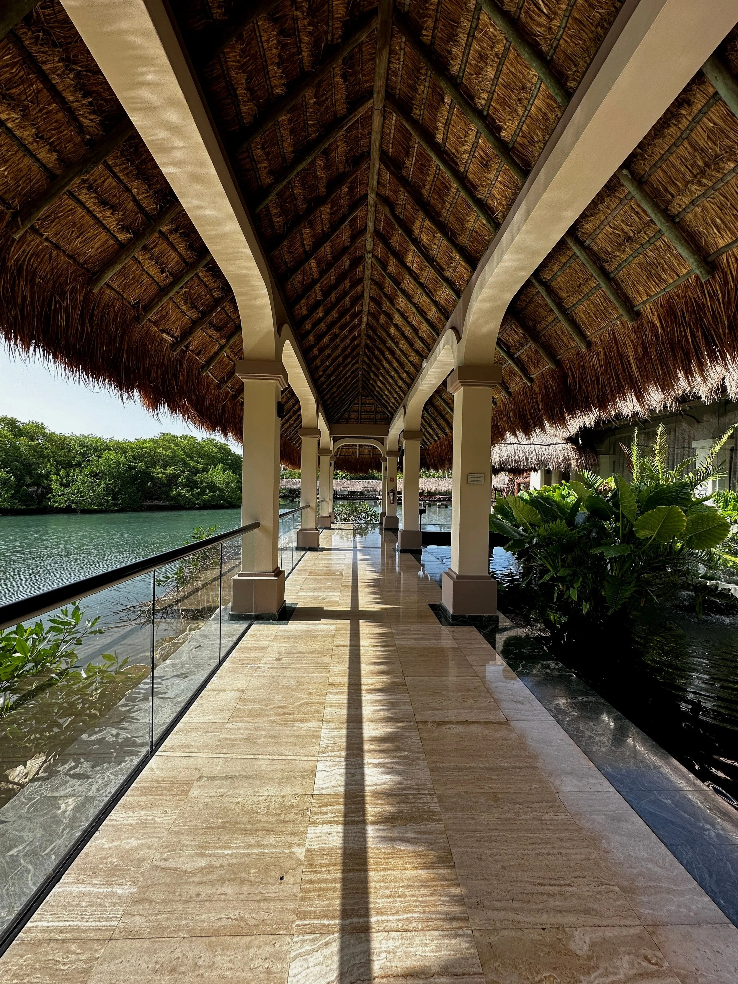 A covered walkway with a thatched roof, beige columns, glass railing, and view of water and green trees on a sunny day.
