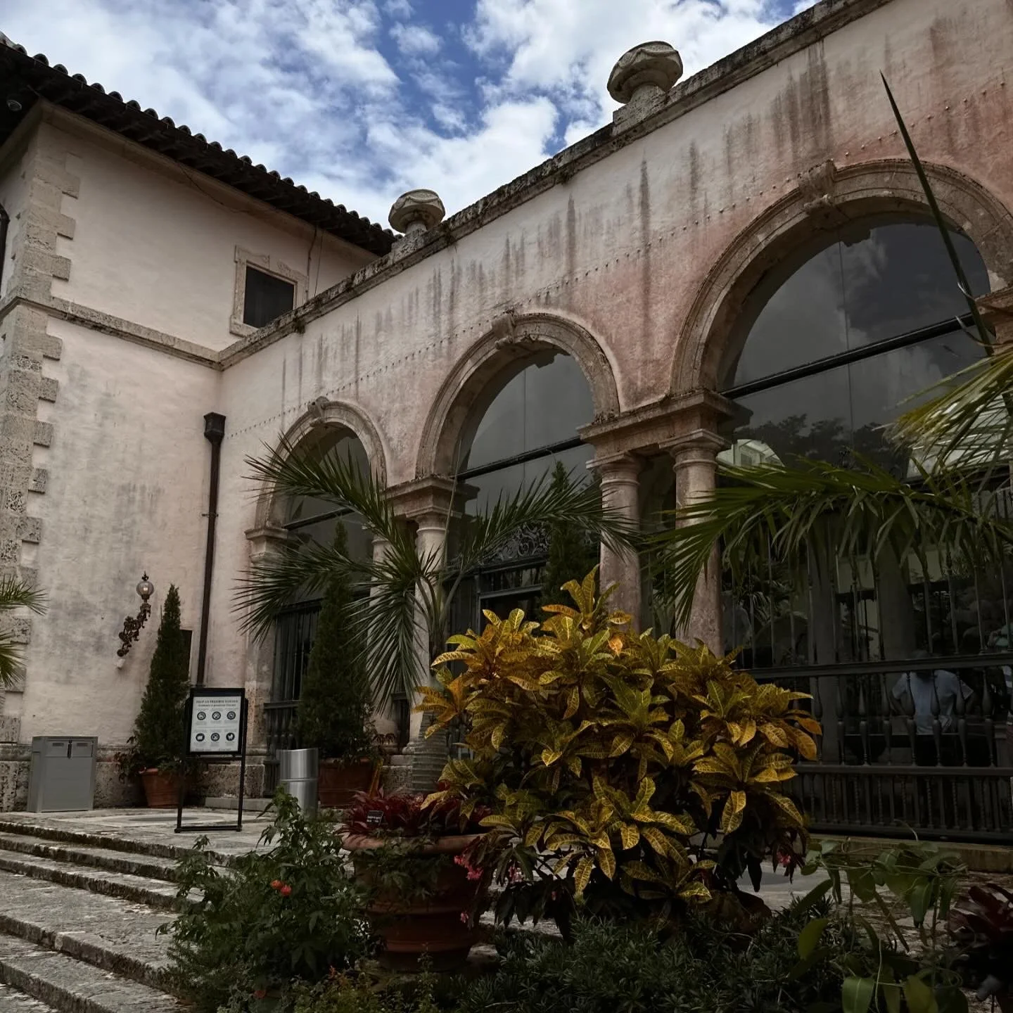 Exterior of an old building with stone arches, windows, and decorative stonework, surrounded by lush plants and potted shrubs, under a partly cloudy sky.