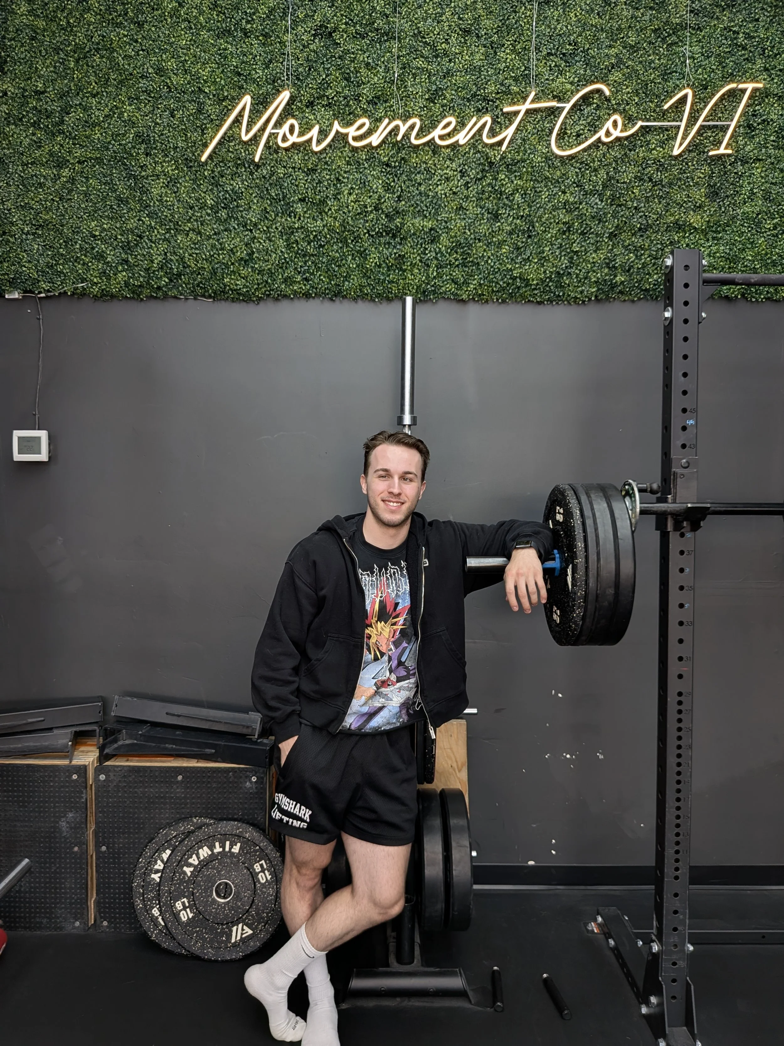 A young man with a smile, standing in a gym, next to weightlifting equipment, with a green hedge wall and neon sign reading 'Movement Co.'