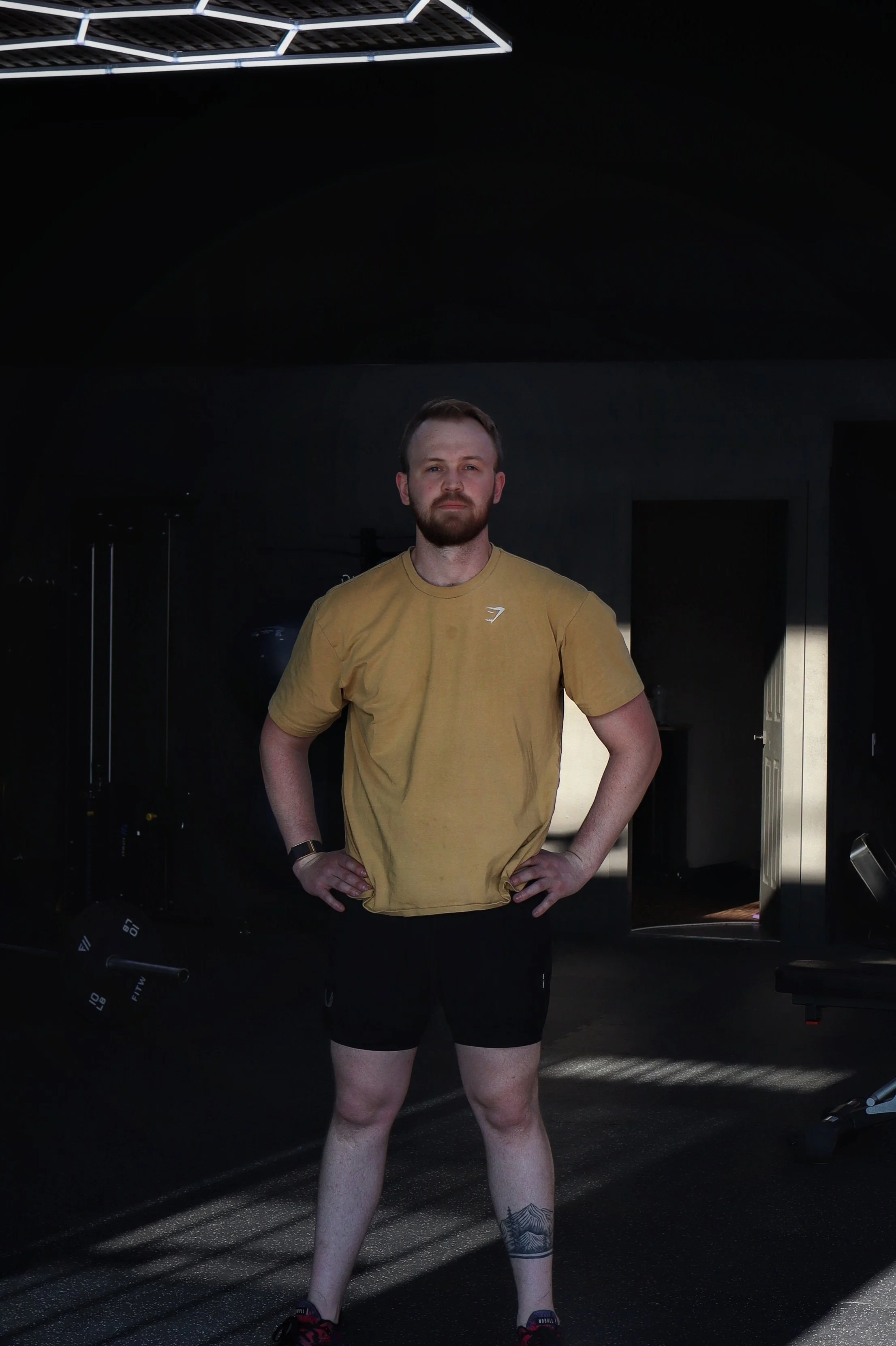 A man with short hair, a beard, and a tattoo on his left calf standing in a gym, wearing a yellow T-shirt, black shorts, and athletic shoes, with a serious expression on his face.