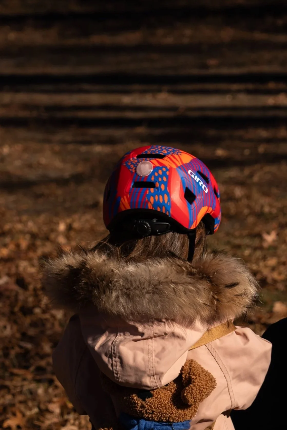 Child wearing a beige winter coat with a fur-lined hood, sitting outdoors on a leaf-covered ground at dusk, with a red and blue bike helmet on their head.