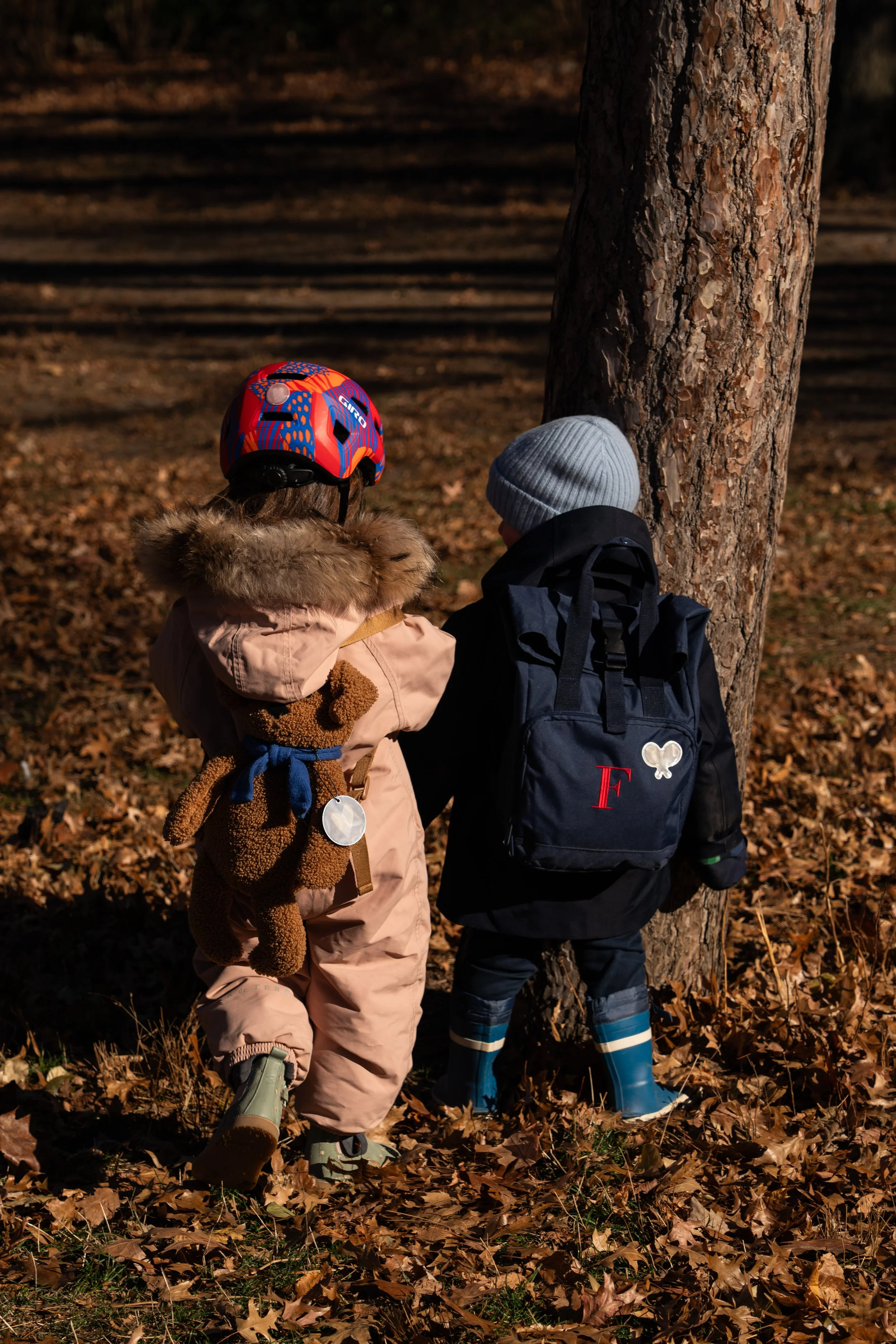 Two children dressed in winter clothing, standing by a tree in a forest with fallen leaves, facing each other and talking.