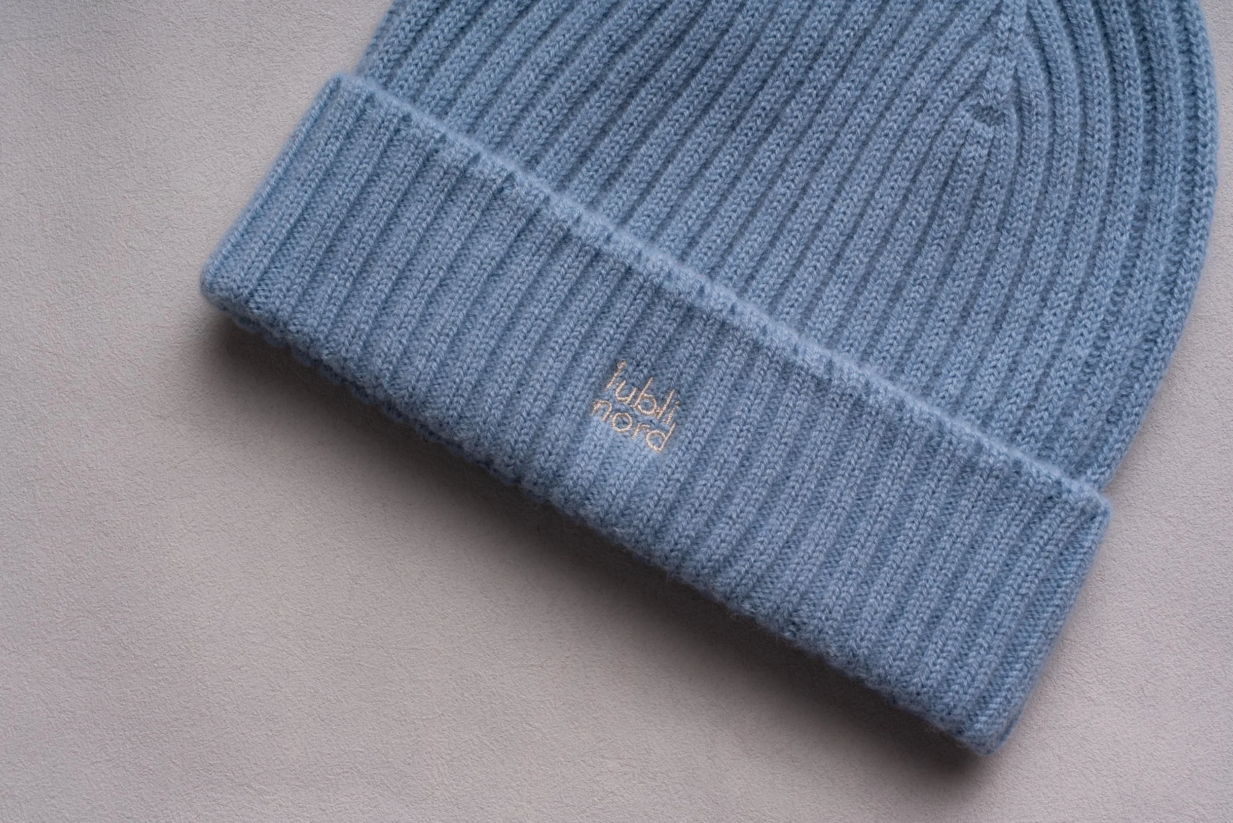 Close-up of a folded light blue ribbed knit beanie with a small metallic logo that reads 'palm hood' on the brim, placed on a light gray textured surface.