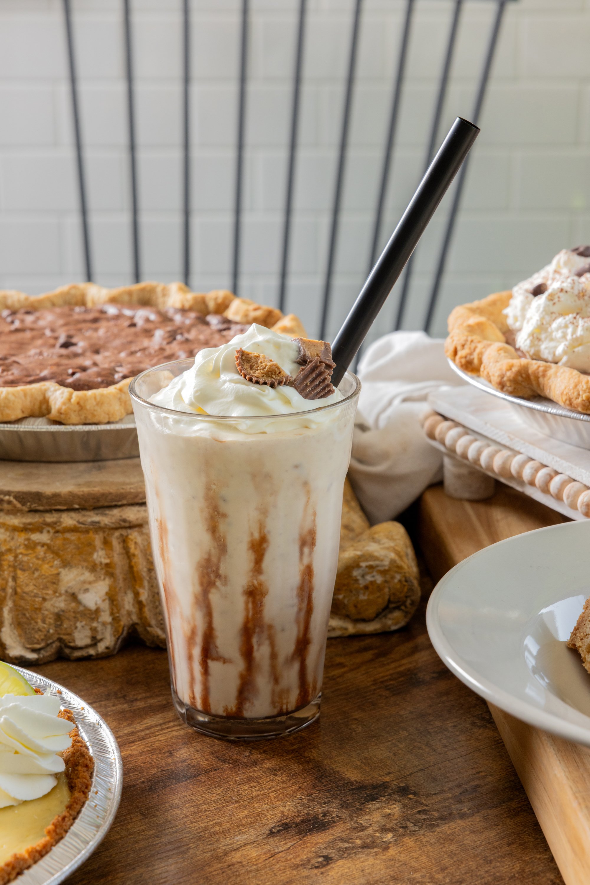 A milkshake topped with whipped cream and a peanut butter cup, surrounded by various pies on a wooden table.