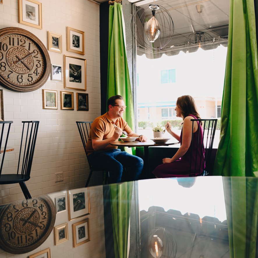 Two people sitting at a small table inside a cafe with green curtains and large wall clock, conversing and eating. Framed photos on white-tiled wall.
