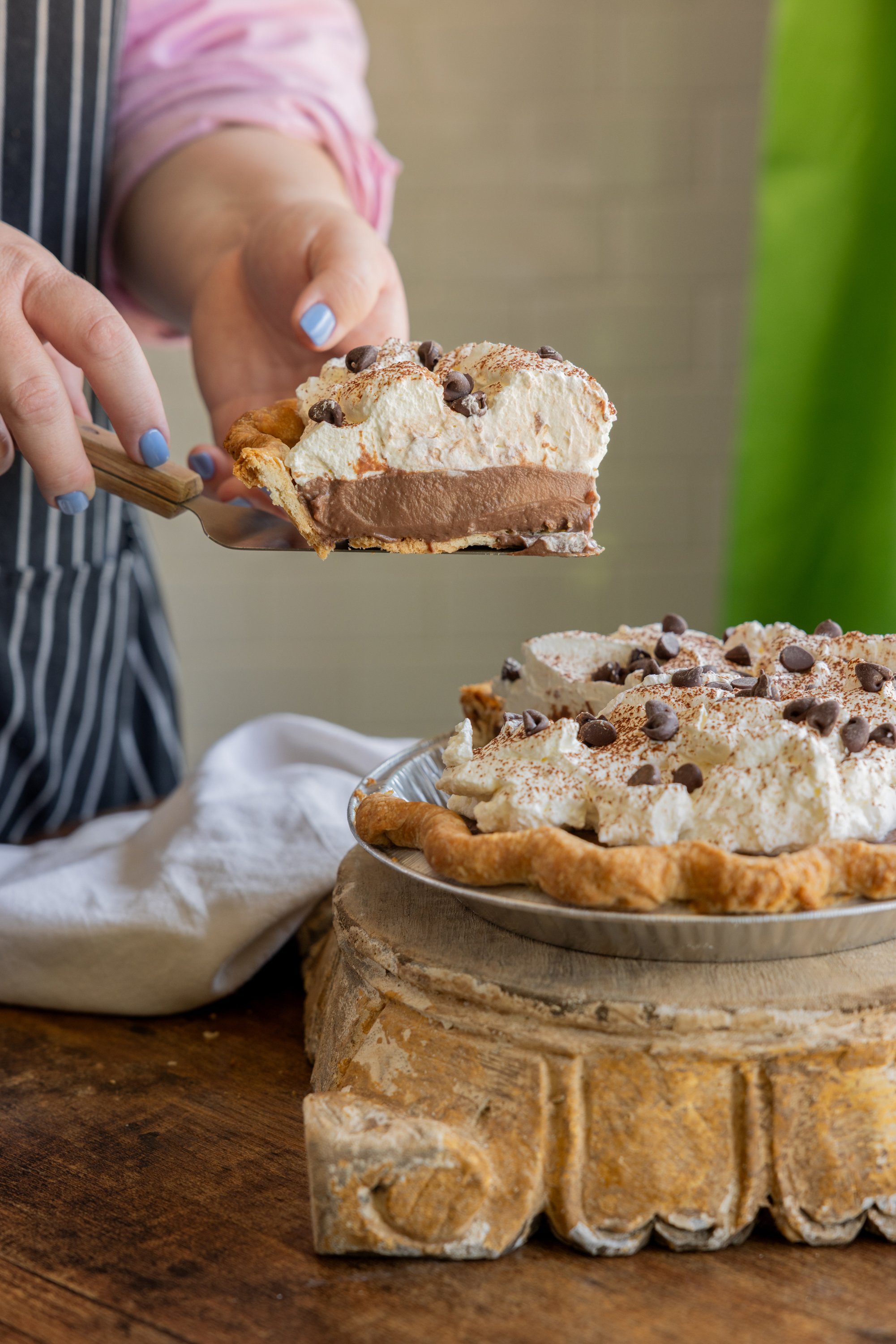 Person serving a slice of chocolate cream pie with whipped topping and chocolate chips from a metal pie dish on a wooden stand.
