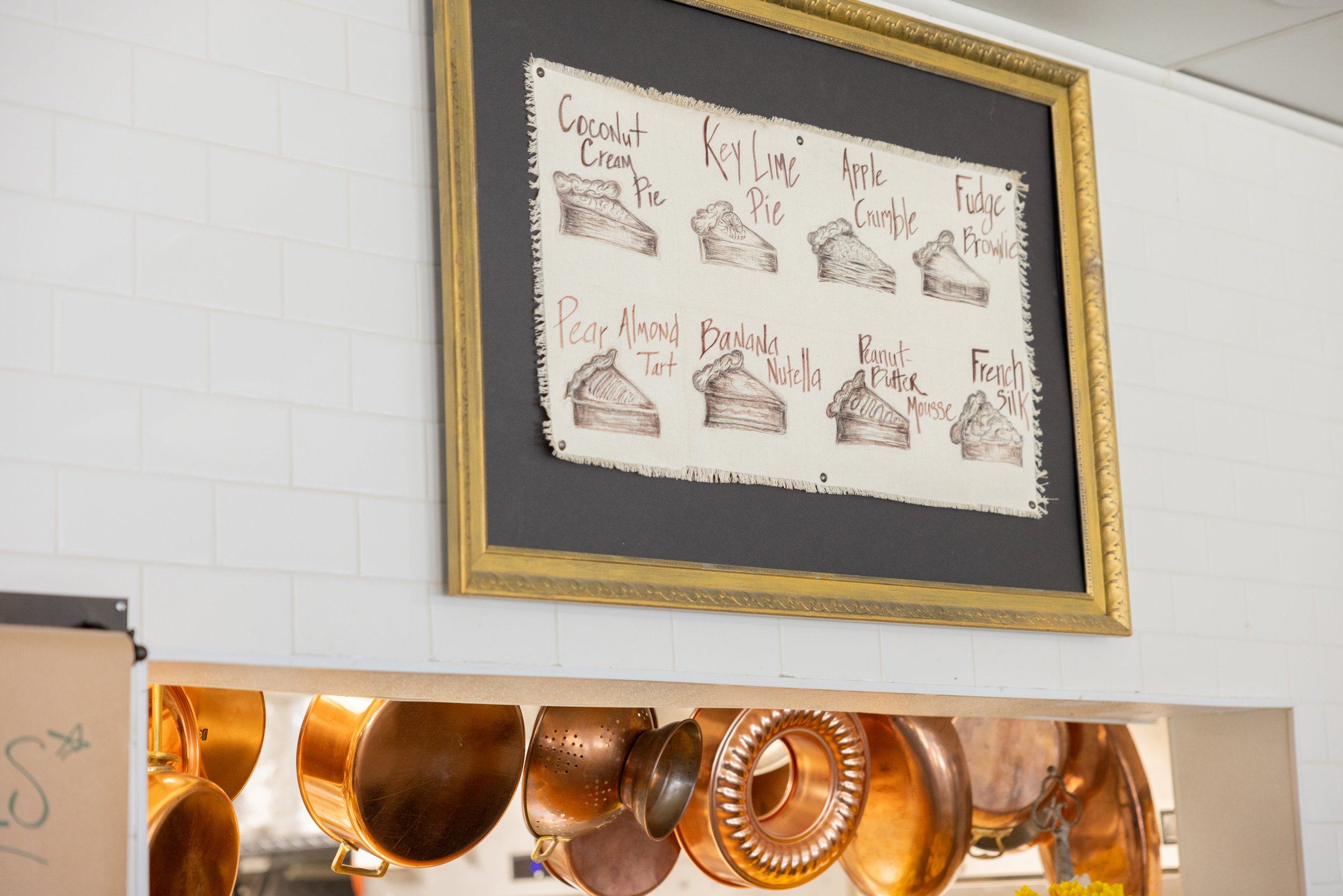 Framed menu with pie illustrations and labels like Coconut Cream Pie, Key Lime Pie, and French Silk above a kitchen area with hanging copper pots and pans.