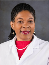 A professional woman wearing a white lab coat, red top, pearl necklace, and earrings, smiling against a gray background.