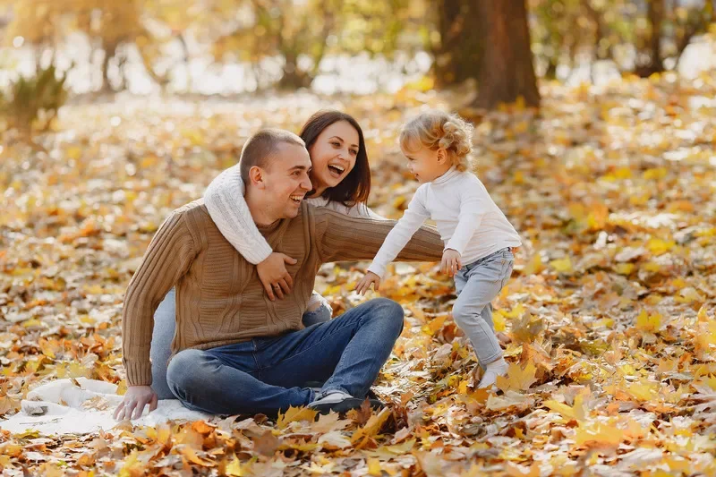 Family of three enjoying a fall day outdoors, sitting on a blanket among fallen leaves, smiling and playing.