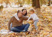 A family of three playing outside in an autumn setting. The father and mother are sitting on the ground, holding hands with their young child and smiling.