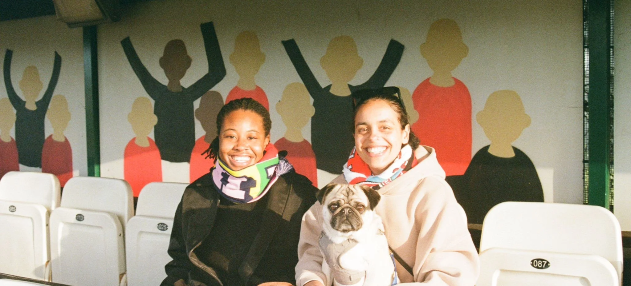 Two women smiling sitting together outdoors with a small dog, with a colorful mural of stylized human figures in the background, and white numbered cafeteria seats in the foreground.