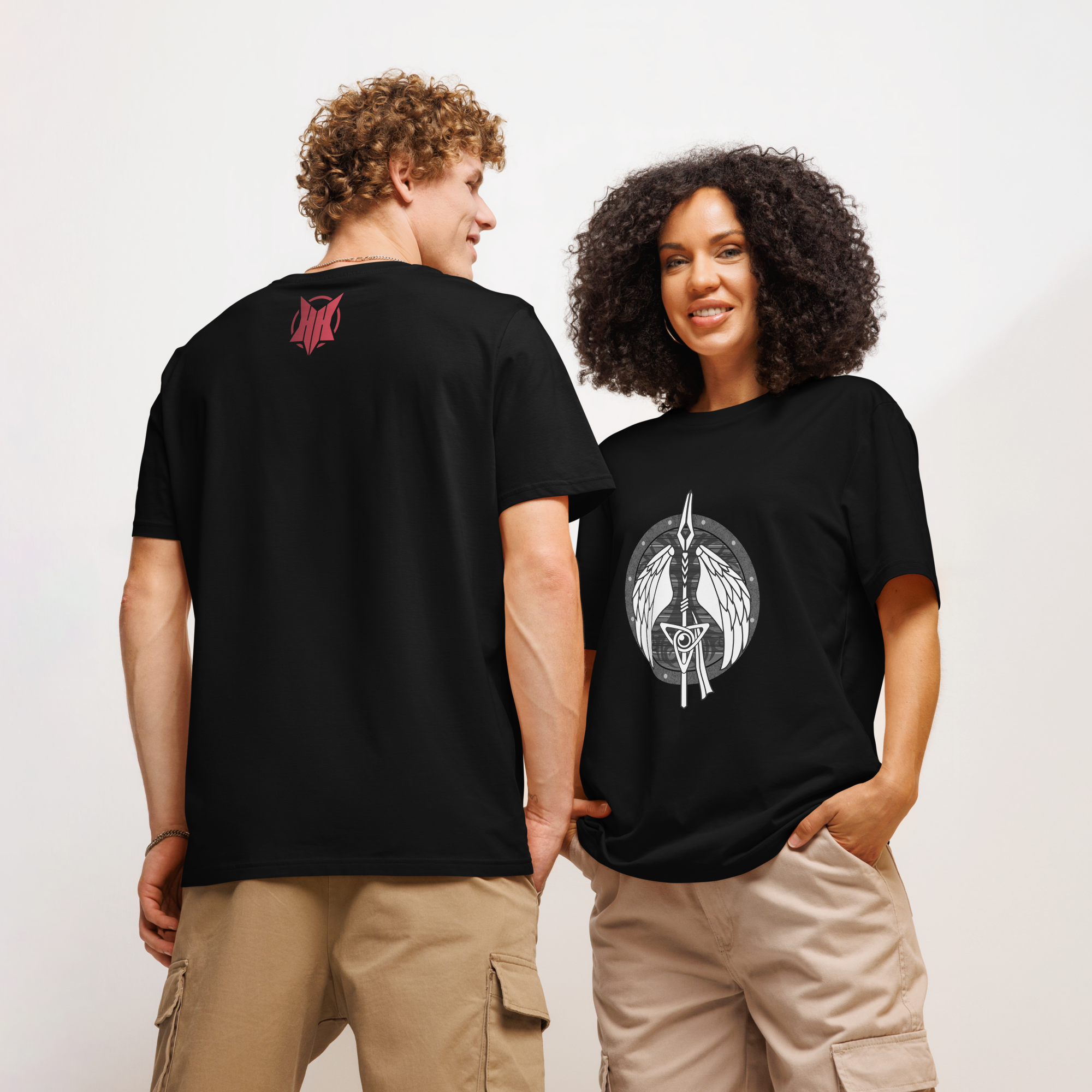 Two people wearing black graphic t-shirts standing against a white background. The person on the right, a woman, has a shirt featuring a sword and wings design. The person on the left, a man, has a smaller design on the upper back of the shirt. Both are wearing beige cargo pants.