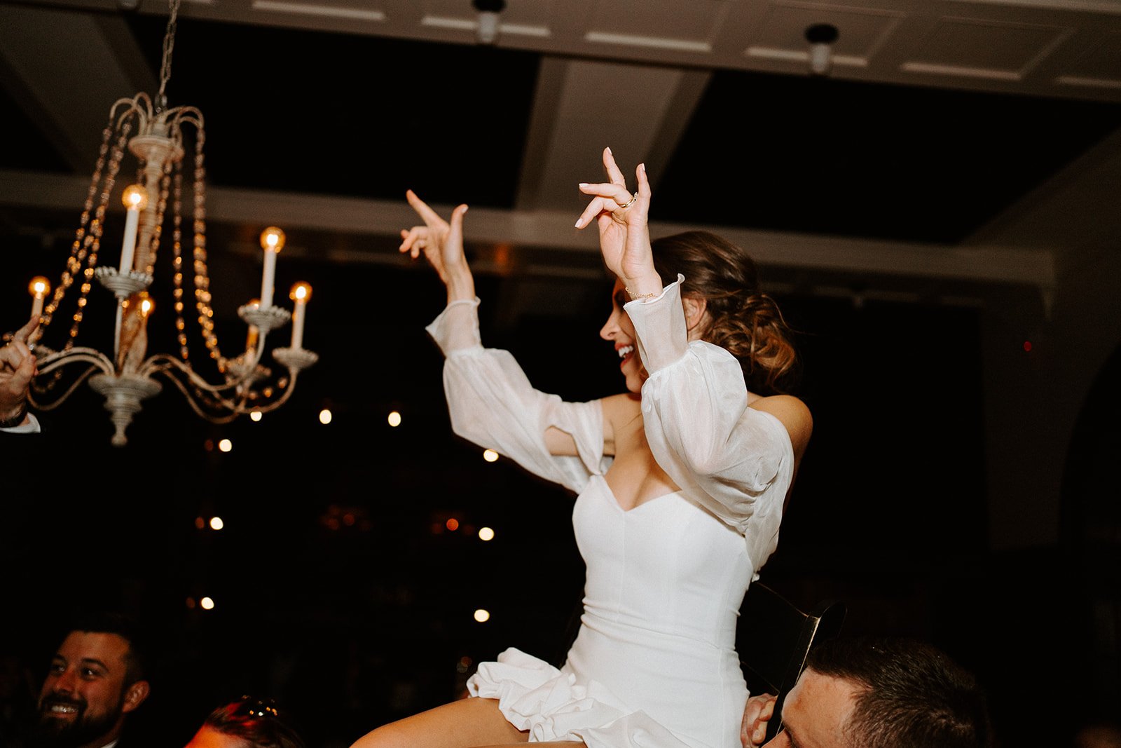 Bride celebrating on shoulders at a wedding reception, with a chandelier overhead.