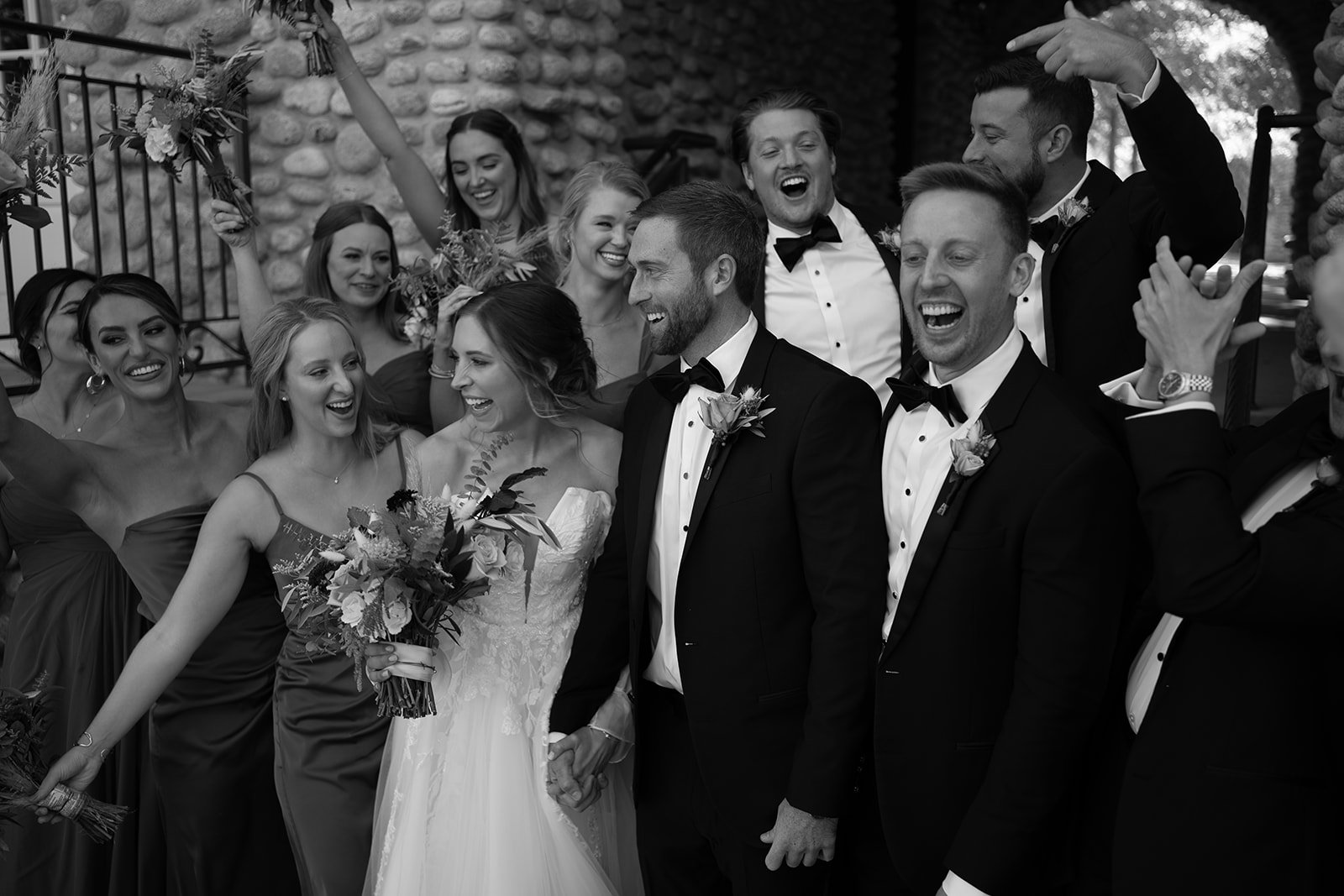 A wedding party celebrating. The bride and groom are in the center, surrounded by bridesmaids in dresses and groomsmen in suits, all smiling and laughing.