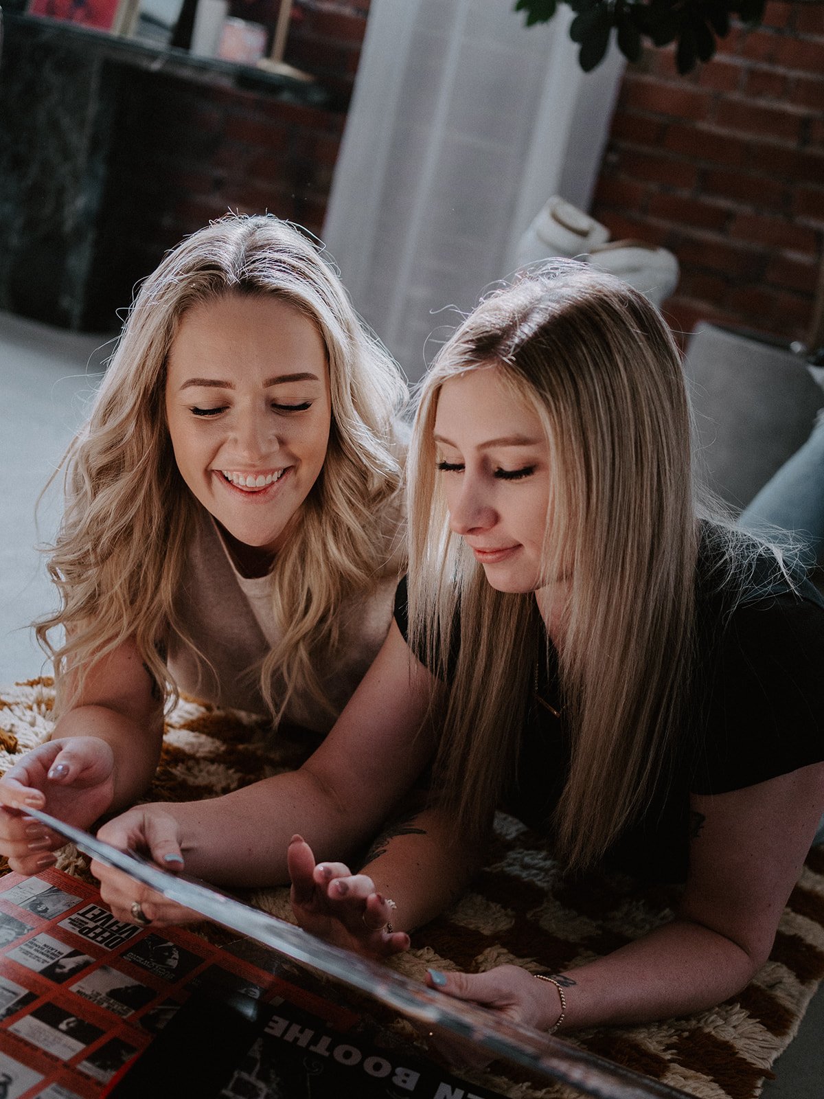Two women lying on a rug, looking at a magazine together, smiling, in a cozy room with brick walls.