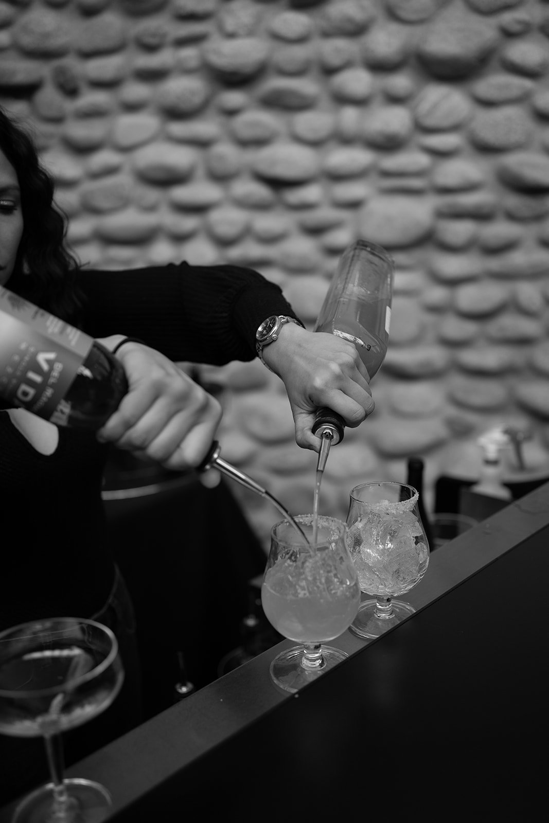 Bartender pouring drinks into glasses with ice, using two bottles at once, against a textured stone wall background.