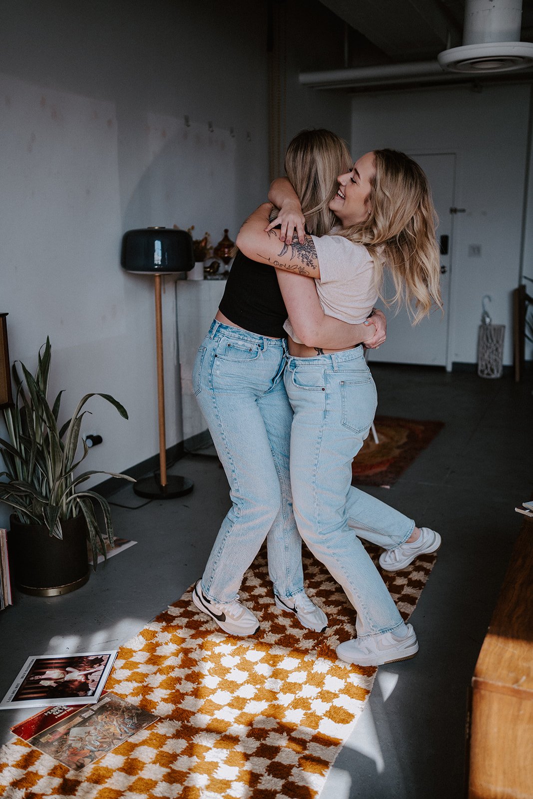 Two people hugging in a room with a checkered rug, indoor plants, and scattered records on the floor. They wear casual outfits with jeans and sneakers, and are smiling in the embrace.